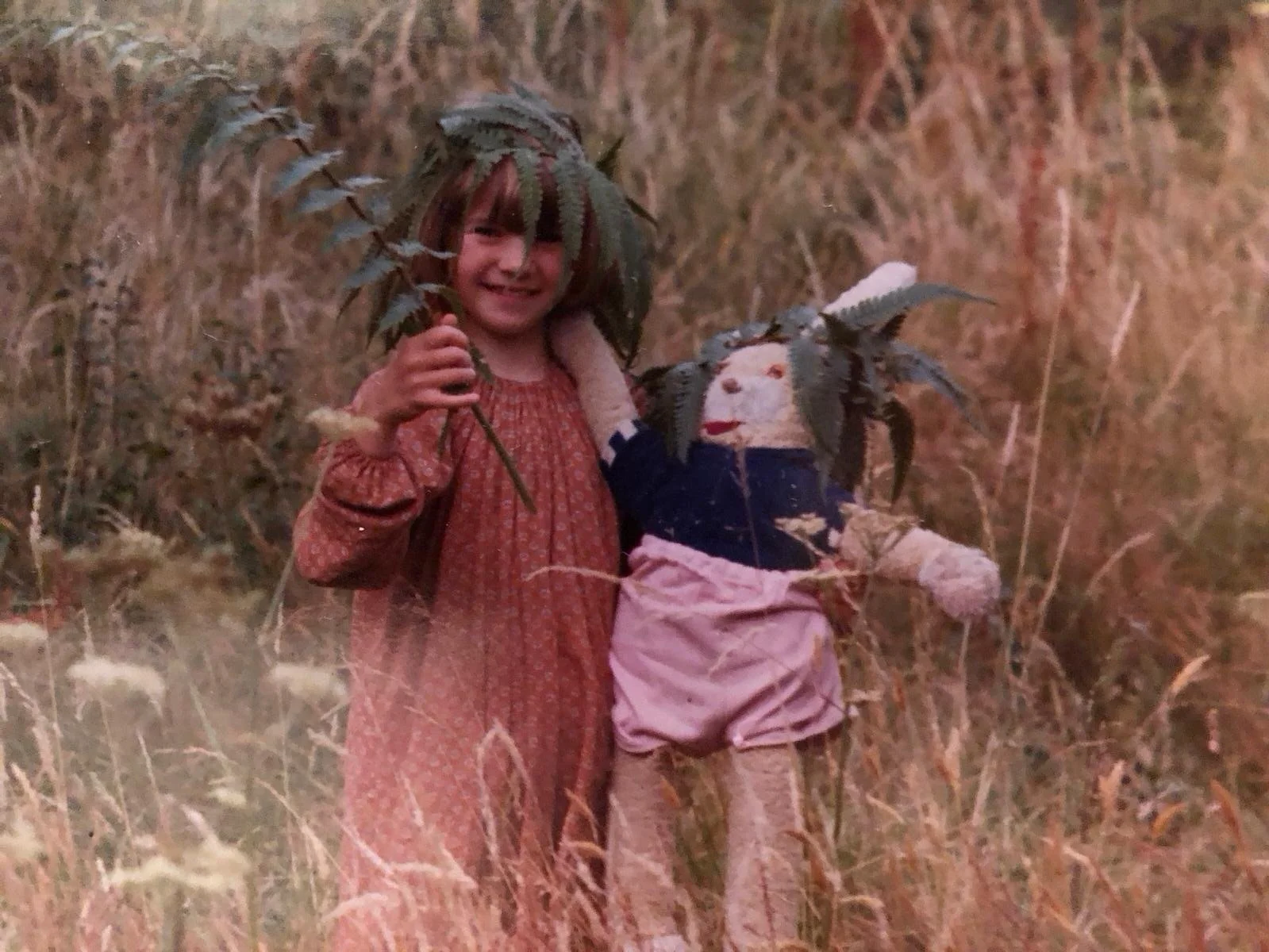A young girl with brown hair, wearing a red dress with long sleeves, is holding a creepy doll dressed in patchwork clothes. The doll has leaves for hair and a painted face, with a dark blue top and pink bottom. The girl and doll are standing in tall 