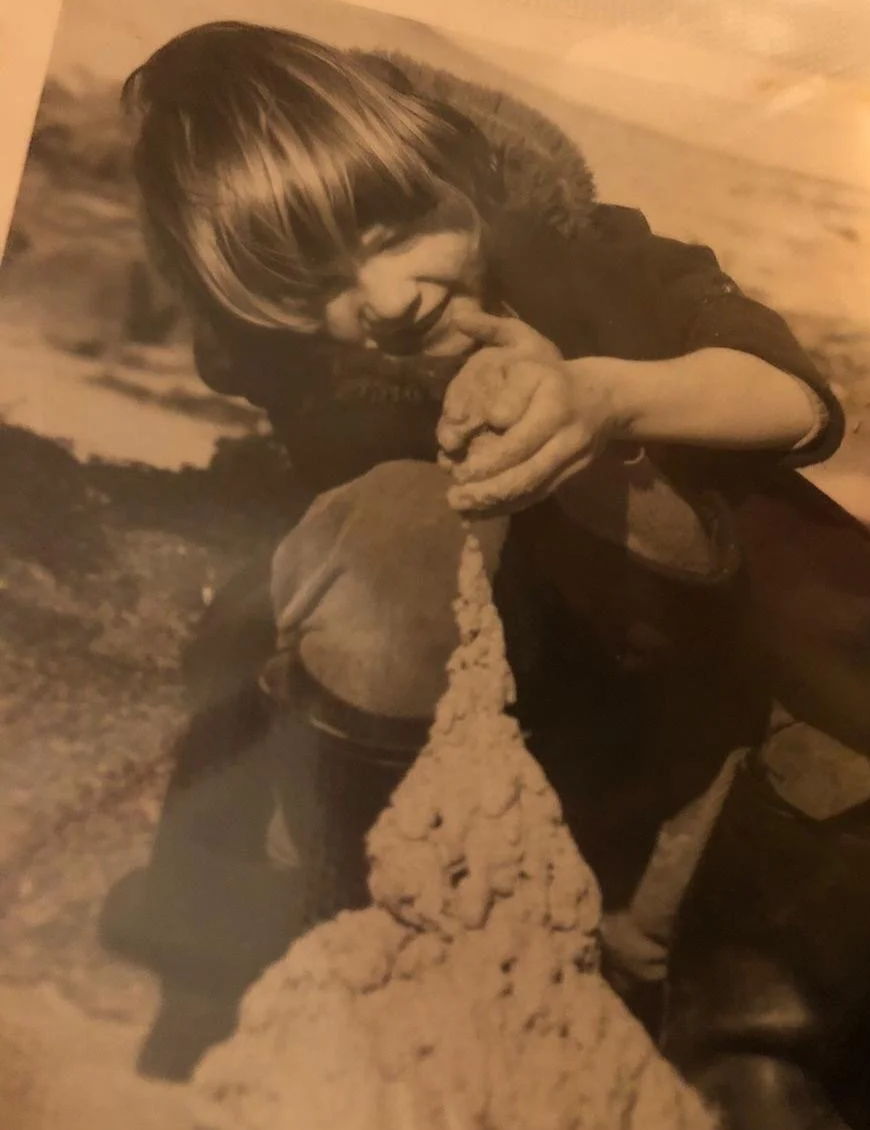 A young child with long hair is playing in the sand and appears to be building a sandcastle with a pointed tower. The child is smiling and focused on their activity.