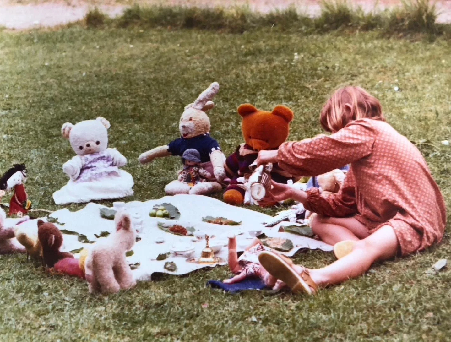A child sitting on grass playing with stuffed animal toys and dolls arranged on a white cloth during an outdoor picnic.