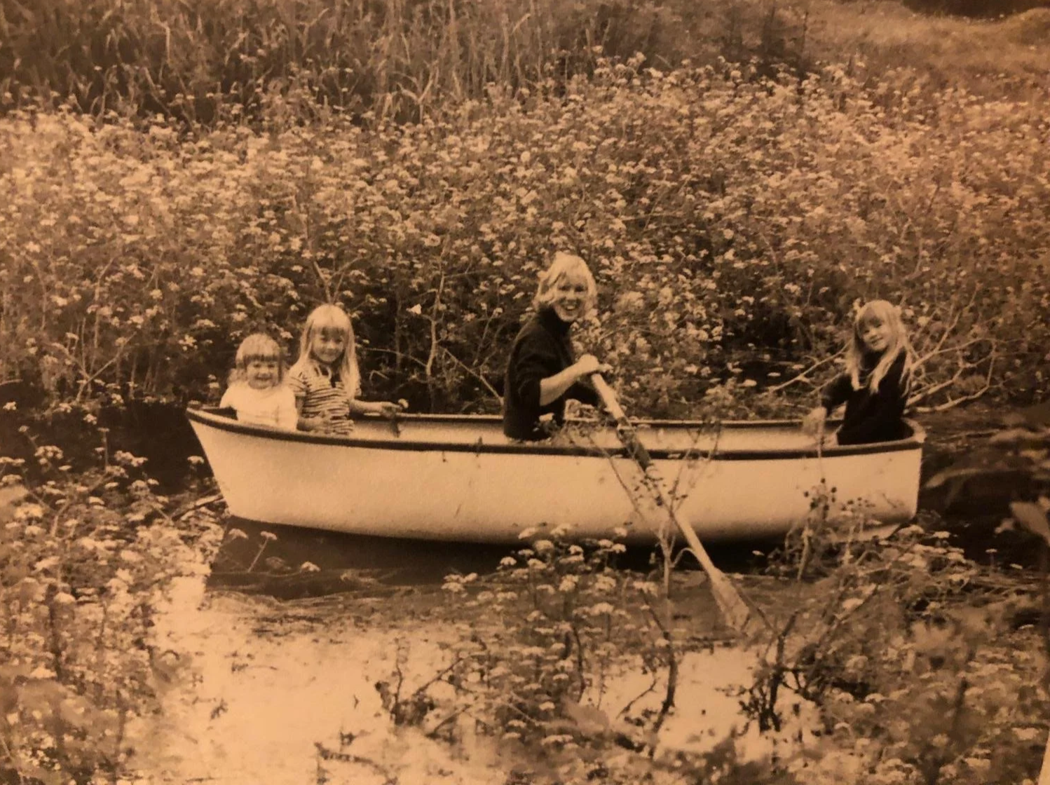 Four children in a small boat on a calm waterway, surrounded by dense bushes and flowers, with one child paddling and the others sitting inside, smiling.