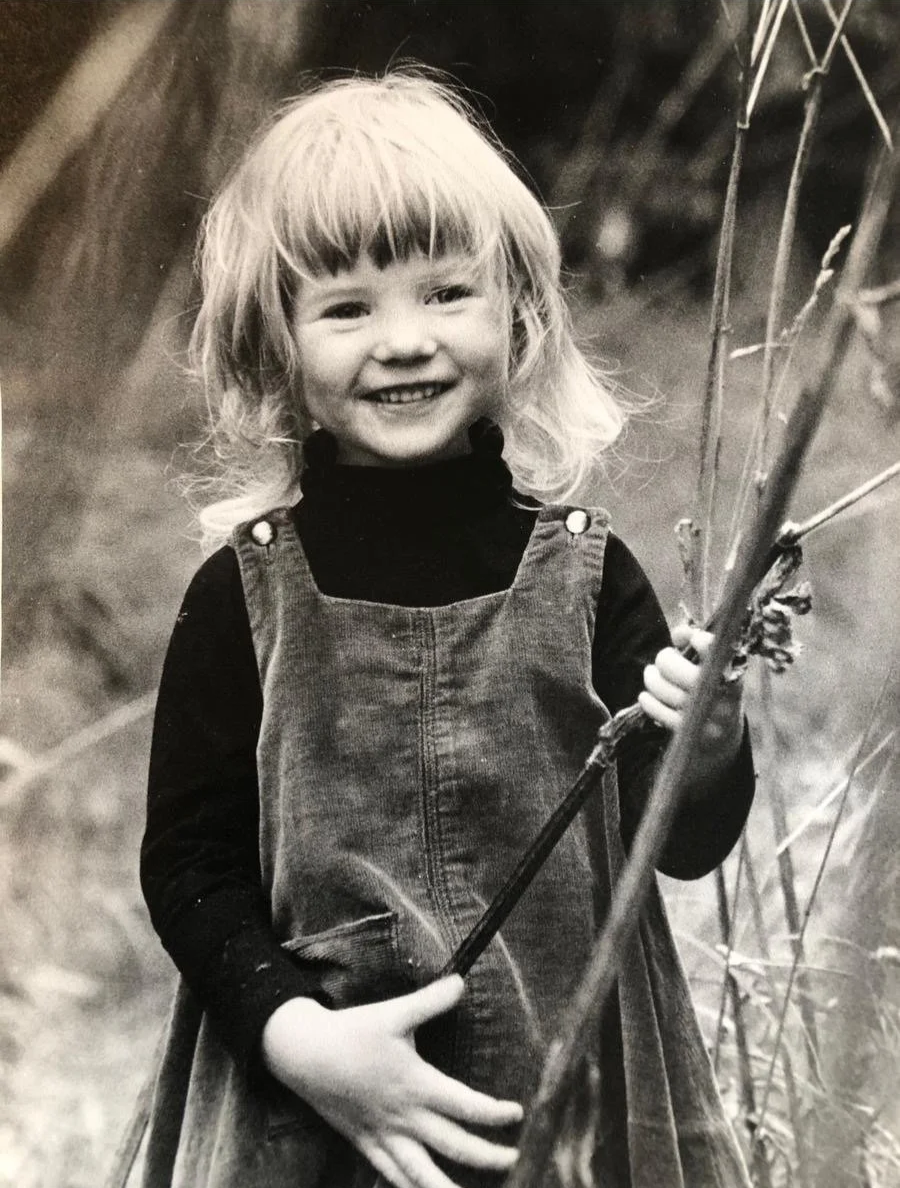 A young girl with blonde hair and bangs smiling while holding a branch, wearing a dark long-sleeved shirt and overalls, outdoors.