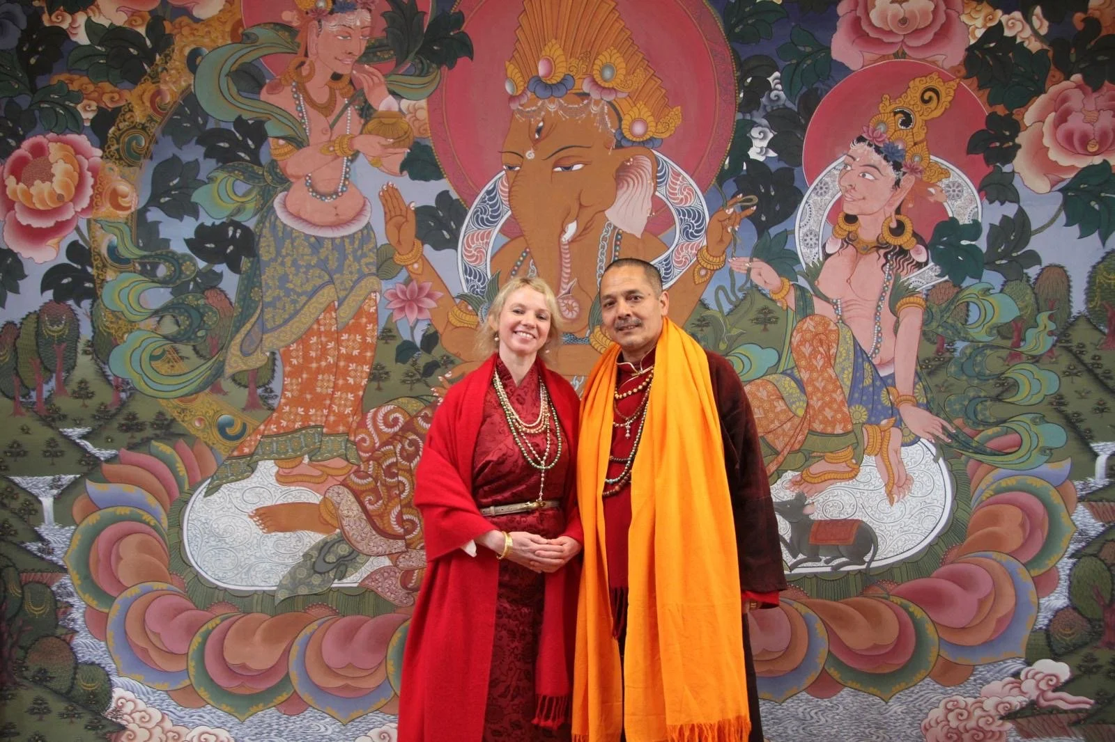 A woman and a man dressed in traditional Indian attire standing in front of a religious mural with Hindu deities, including Ganesha, painted with vibrant colors and intricate designs.