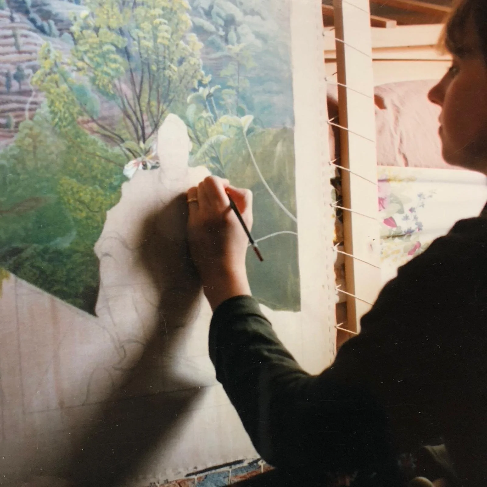 A person is painting a large outdoor landscape on a canvas, featuring trees, plants, and a mountain in the background.