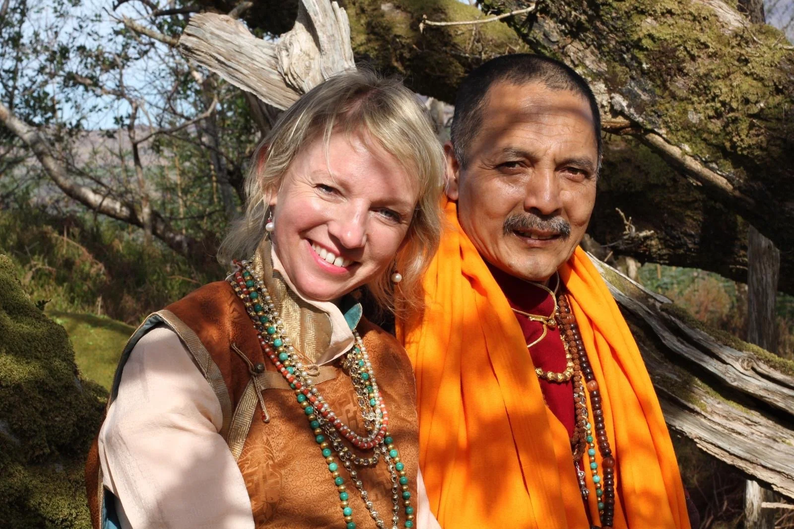 A woman with blonde hair and a man with dark hair and a mustache, both wearing colorful traditional clothing and necklaces, standing outdoors near a large fallen tree.