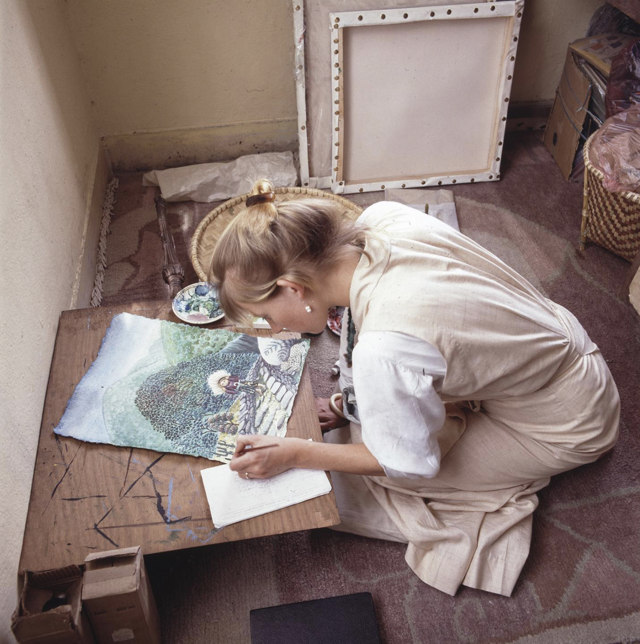 A woman kneeling on the floor, working on a colorful artwork of a tree and a character, with sketchbook and art supplies nearby, in a room with a brown carpet and a beige wall.