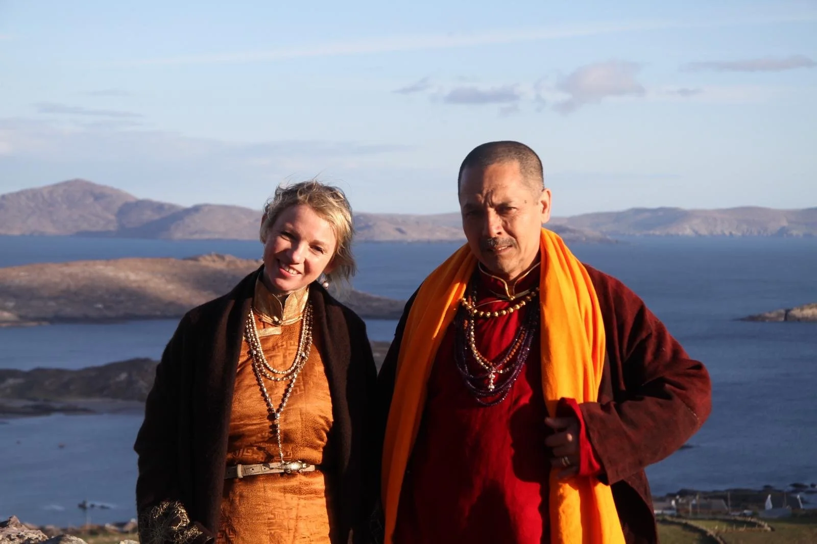 Two individuals dressed in traditional clothing standing outdoors with a lake and mountains in the background.
