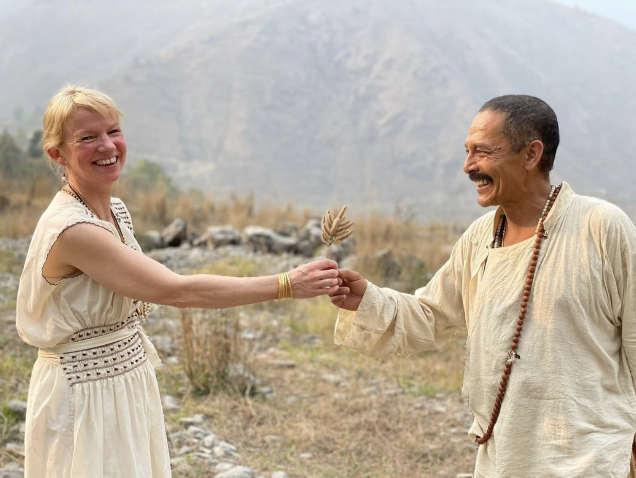 A woman and a man, both smiling and laughing, exchange a small bundle or object outdoors in a dry, mountainous landscape.
