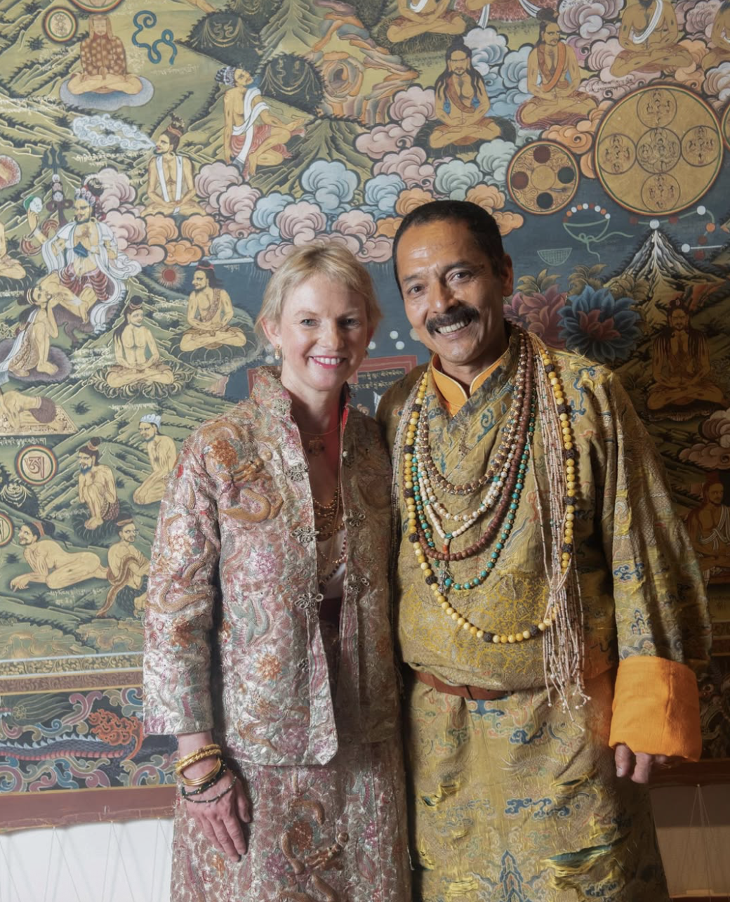A woman and a man dressed in traditional clothing standing in front of a detailed, colorful Buddhist mural depicting monks, deities, and religious symbols.