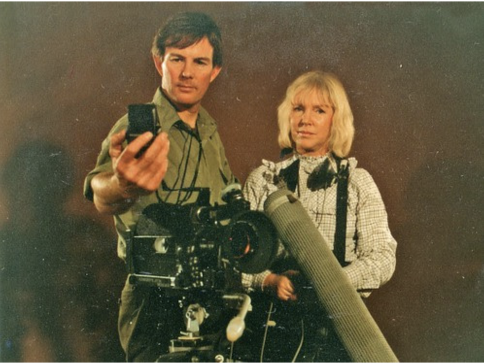 A man with brown hair and a woman with blonde hair pose with film production equipment including a camera and a microphone, against a plain background.