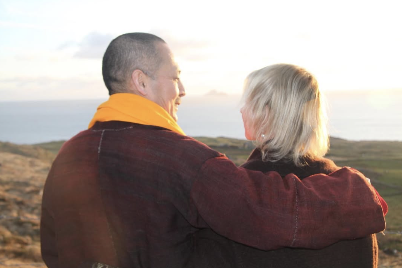A couple embracing outdoors during sunset, overlooking a landscape with water in the distance.