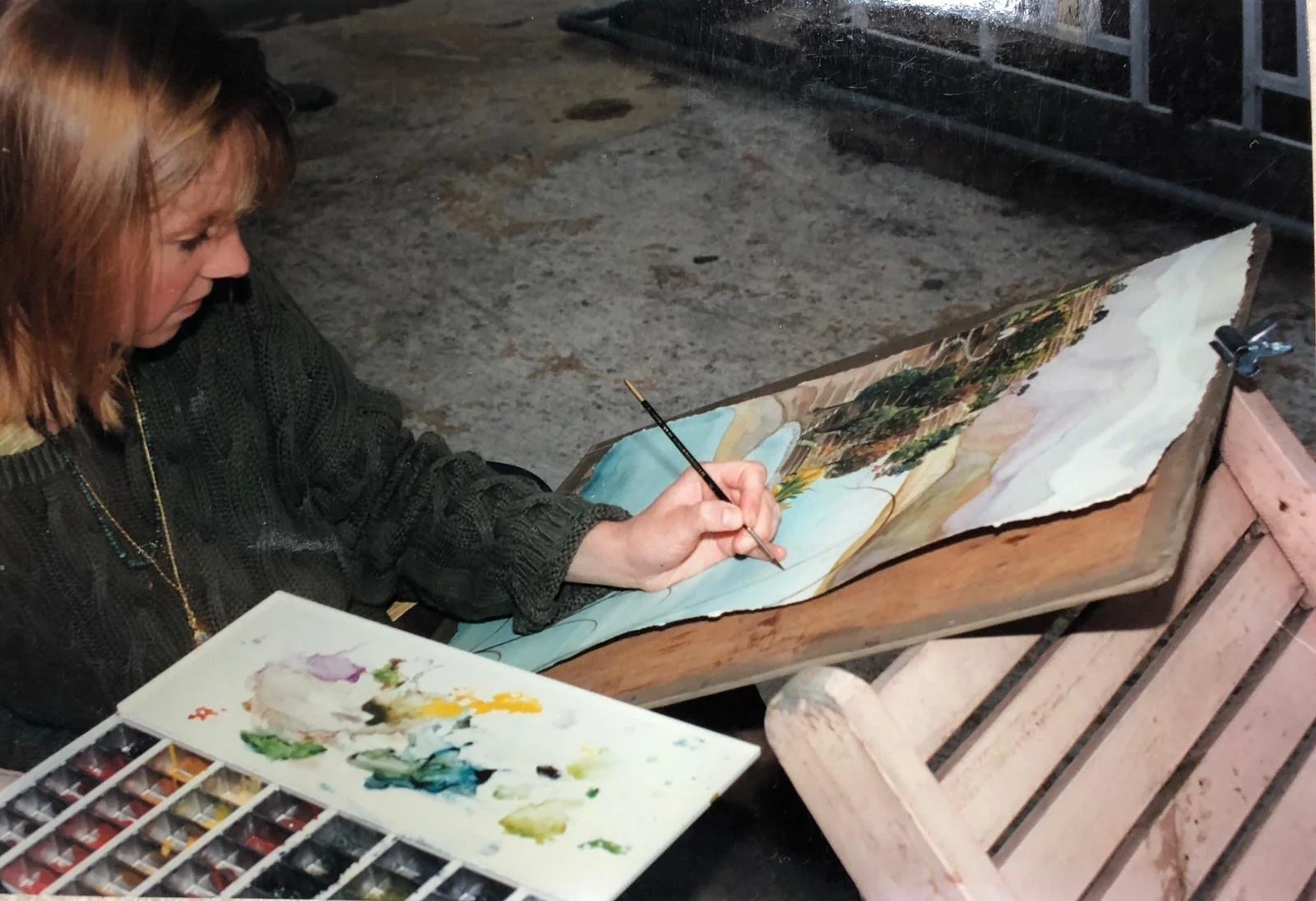 Woman painting a winter landscape on a wooden board outdoors, with a palette of watercolor paints nearby.