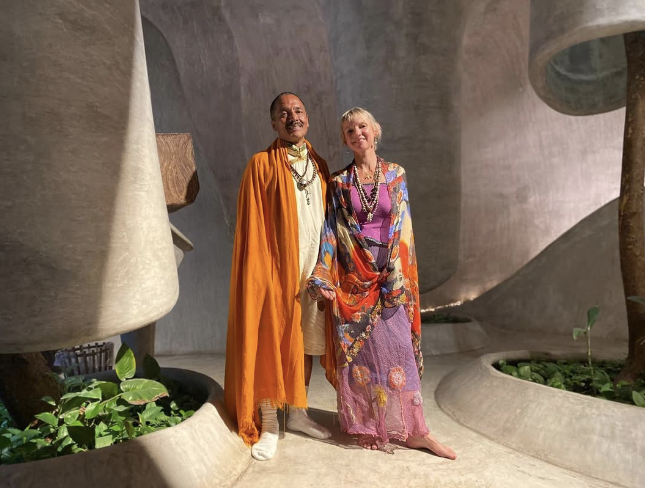 Two people, a man dressed as a Hindu monk in orange robes and a woman in colorful, floral-patterned clothing, standing and smiling together in a modern, sculptural interior setting.