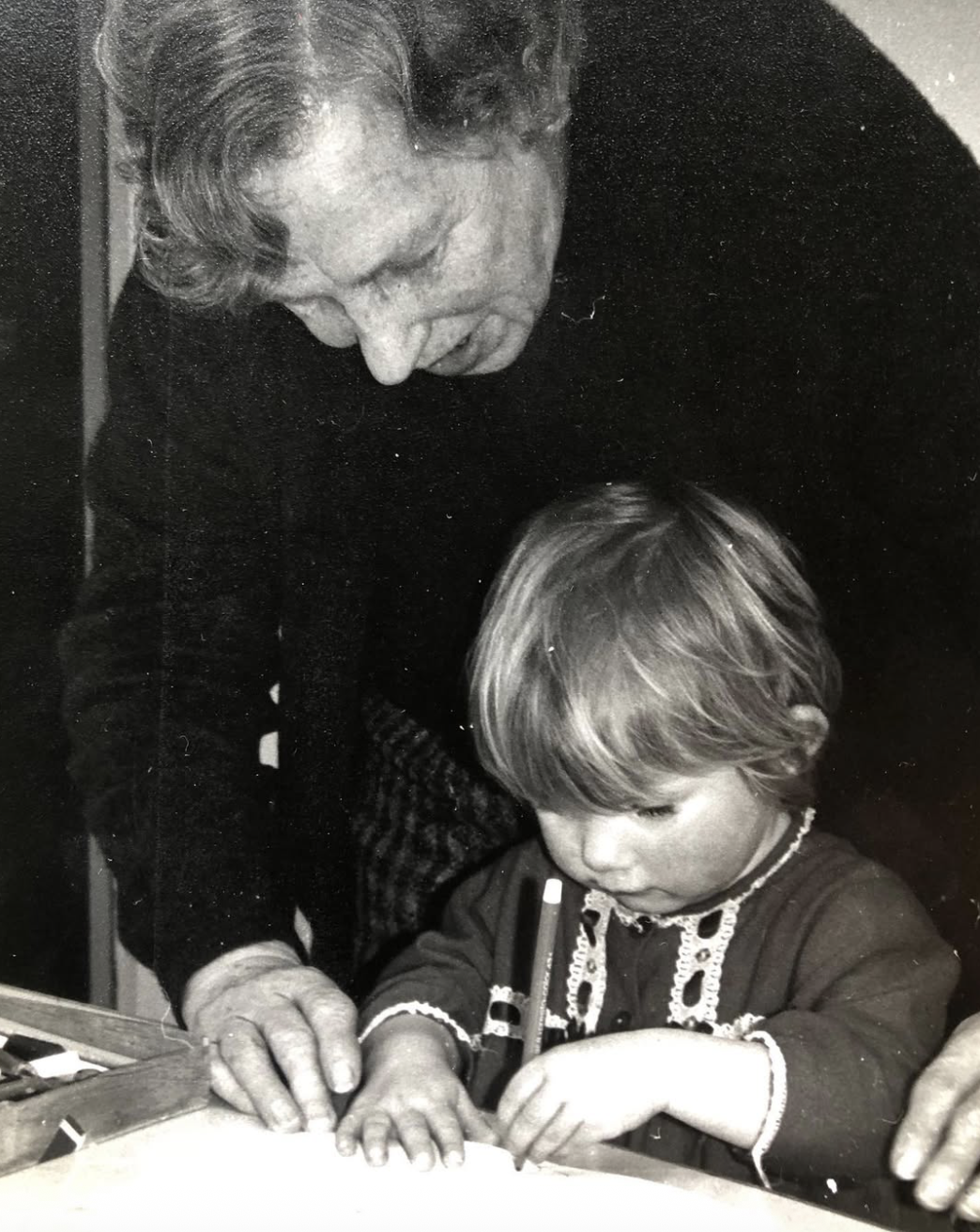An older woman teaching or guiding a young child who is drawing or writing with a pencil on a piece of paper.