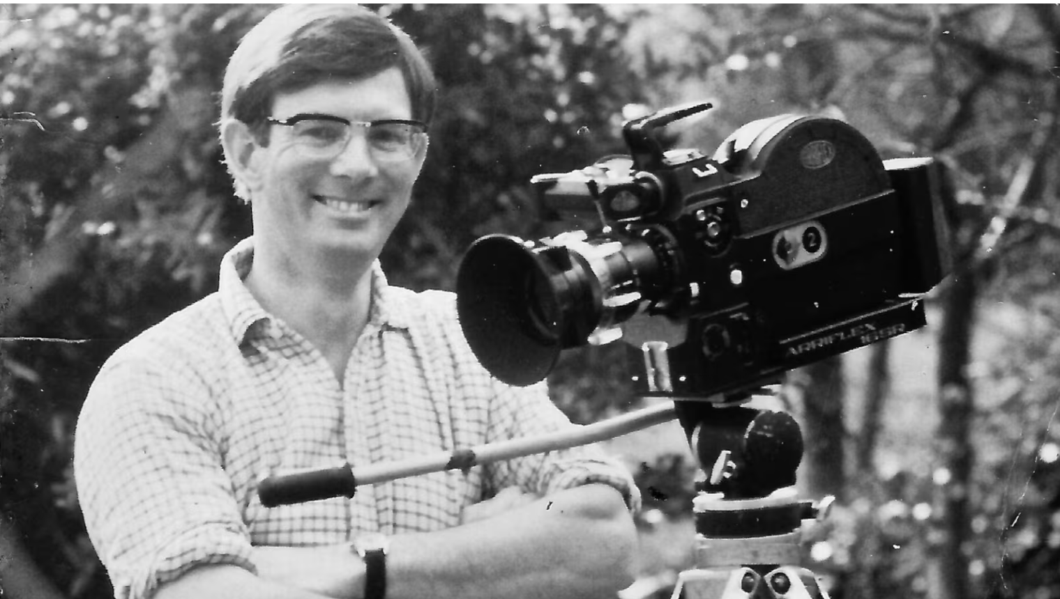 A man smiling and looking at the camera, standing outdoors, operating a film camera on a tripod.