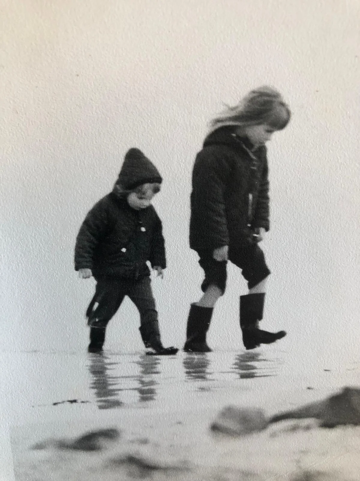 A black-and-white photo of a woman and a boy walking in shallow water. They are both dressed in warm clothing and boots, with the woman slightly ahead and looking down, and the boy following and looking down at the water.