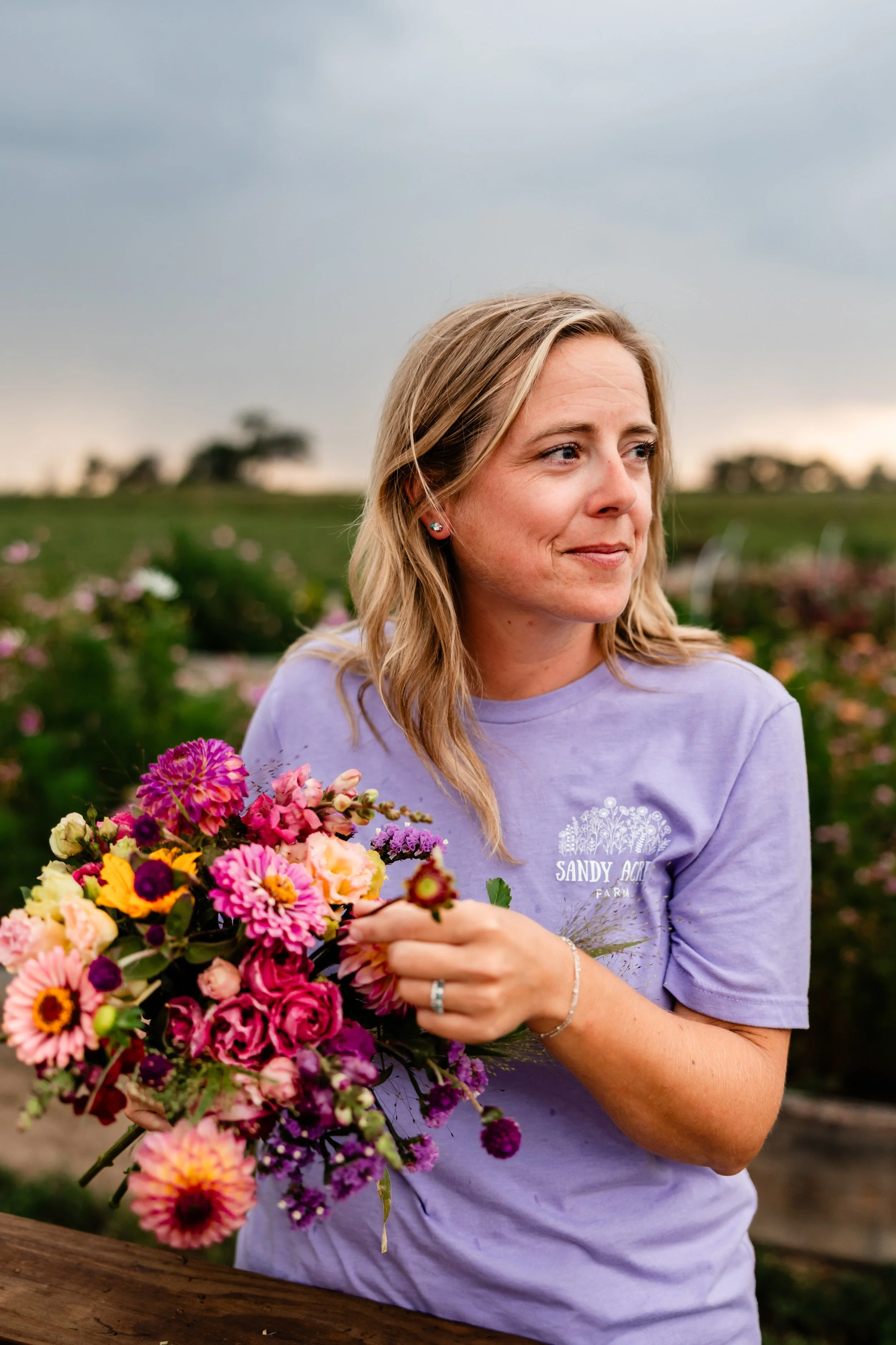 A woman with blonde hair, wearing a purple t-shirt that reads 'Sandy Acres Farm,' is holding a colorful bouquet of flowers outdoors, with a field of flowers and a cloudy sky in the background.