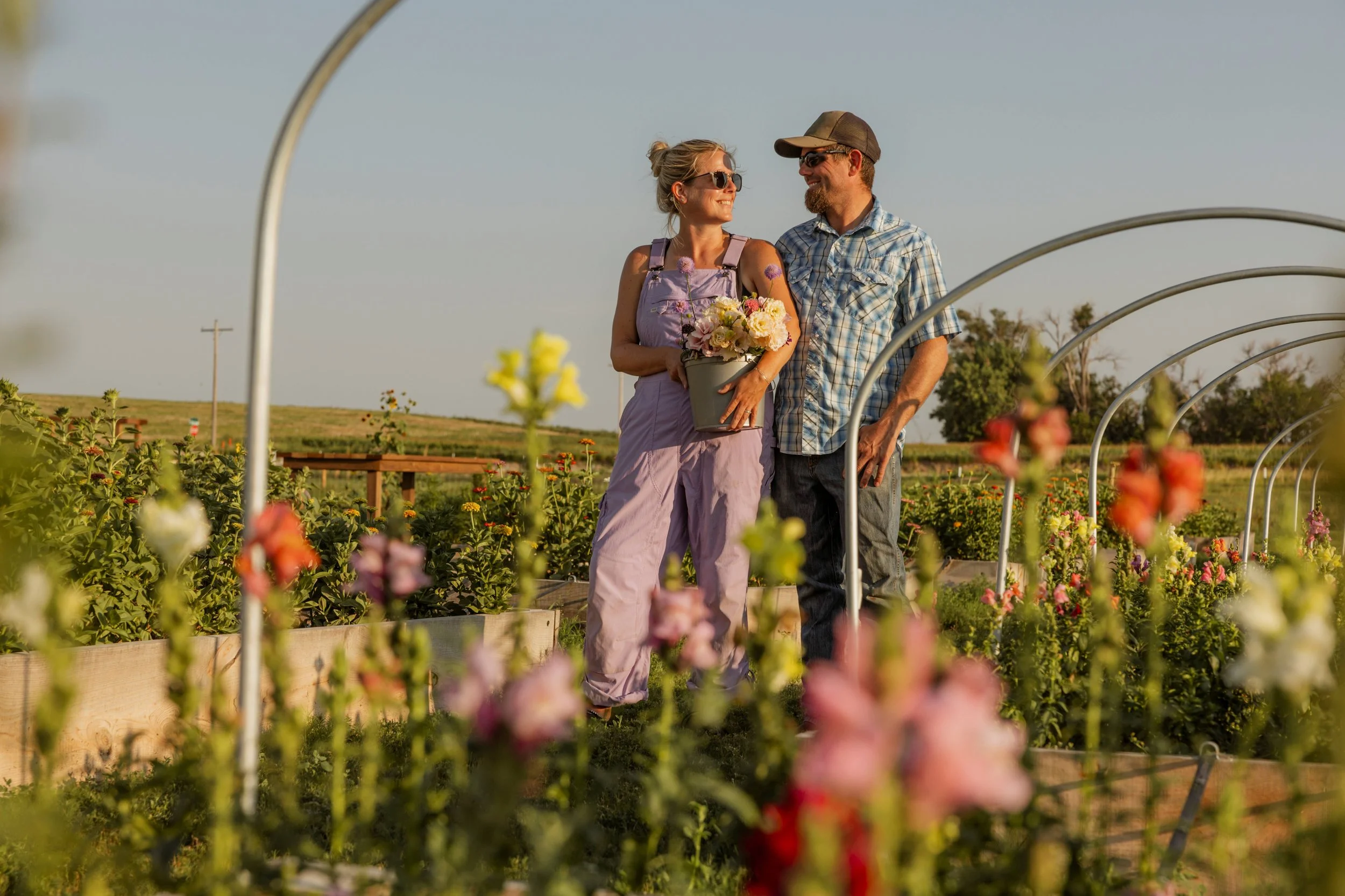 A couple walking through a flower garden, smiling and looking at each other, with the woman holding a pot of flowers, during sunset.