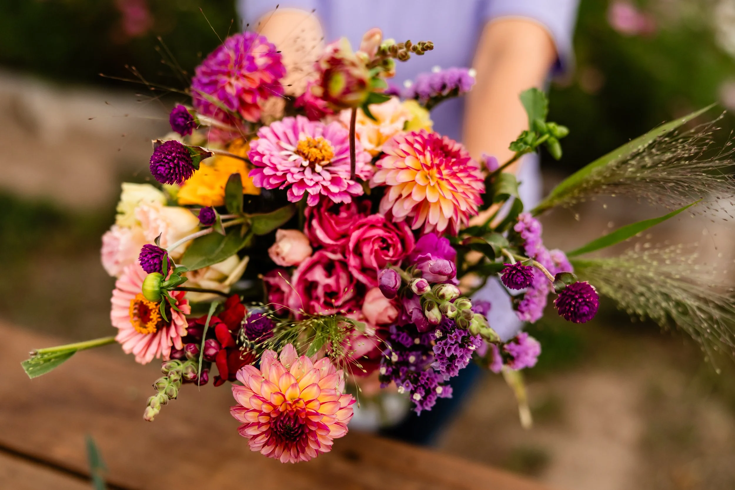 A colorful bouquet of various pink, purple, yellow, and orange flowers, including dahlias, roses, and other blooms, held by a person in a purple shirt.