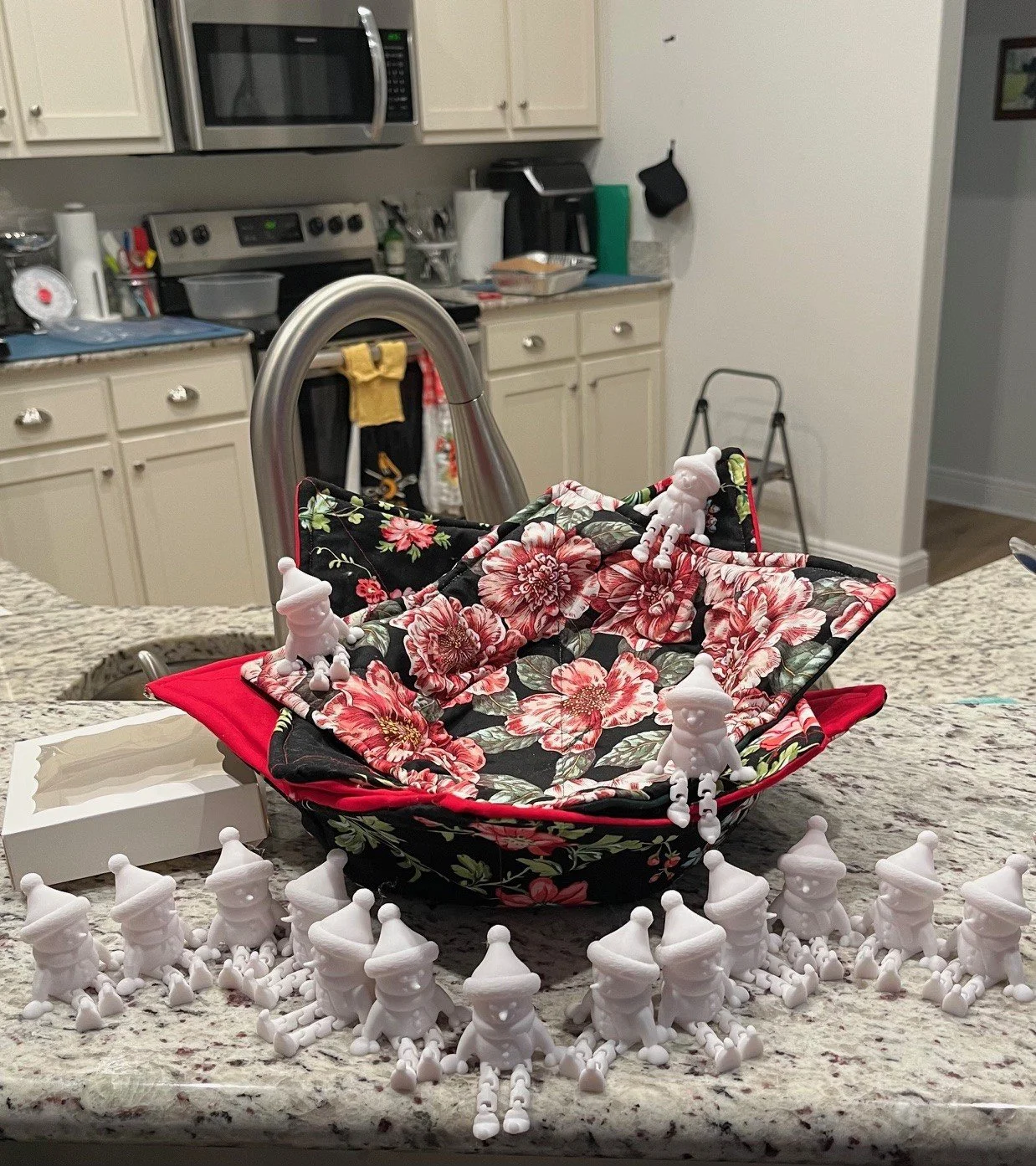 A basket of different size bowl cozies using a floral cotton fabric interior and red exterior, surrounded by small white 3D articulated snowmen figurines on a kitchen countertop.