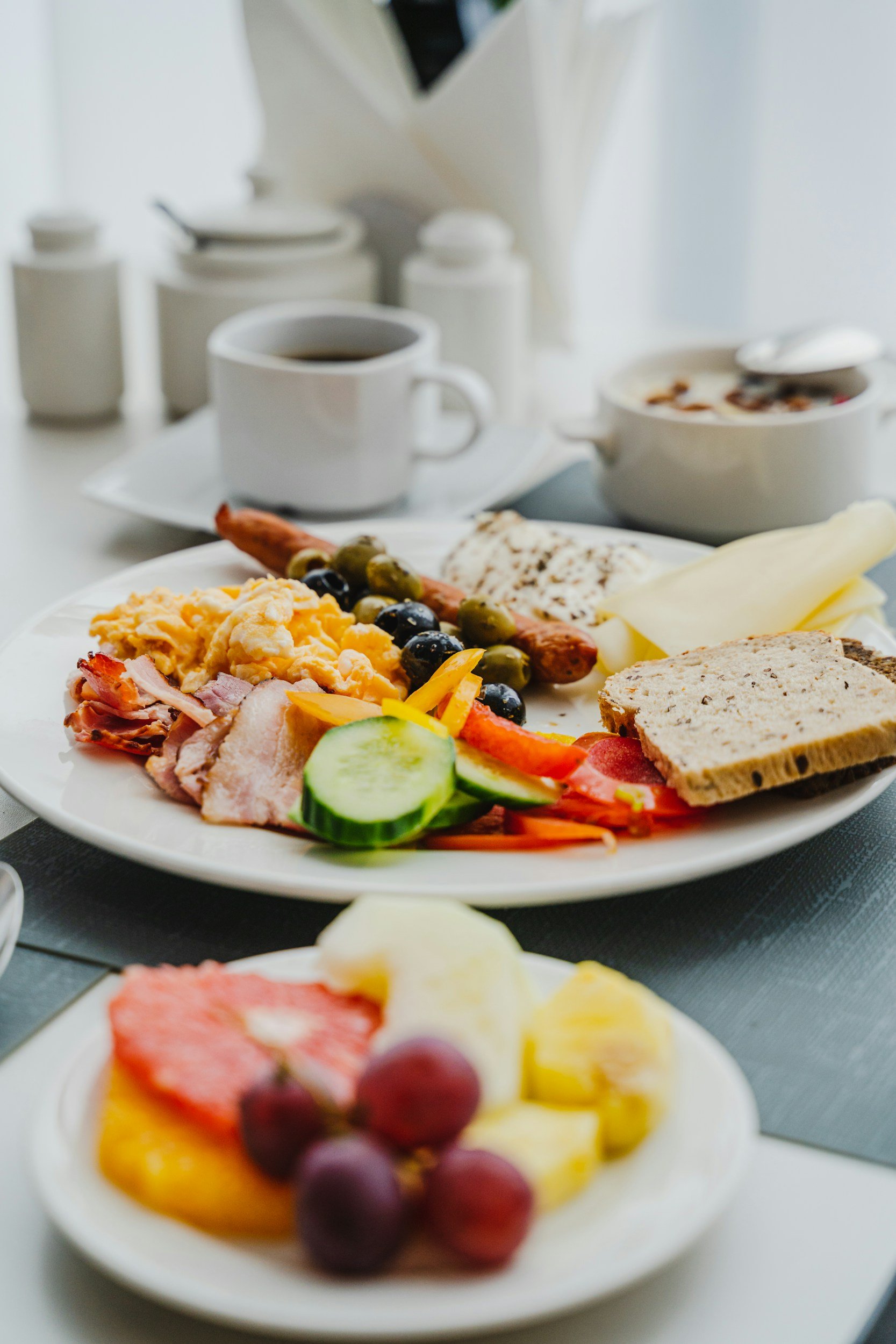 Close-up of a breakfast spread featuring assorted fruits, scrambled eggs, slices of bread, and a cup of coffee on a white table with a bowl of cereal and sugar containers in the background.