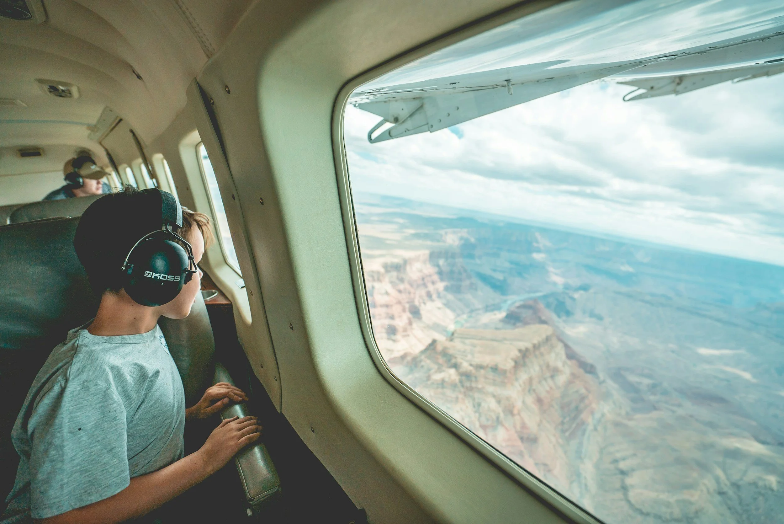 A young boy wearing large headphones looks out the window of an airplane at the landscape below, which includes canyons and mountains, during daytime.