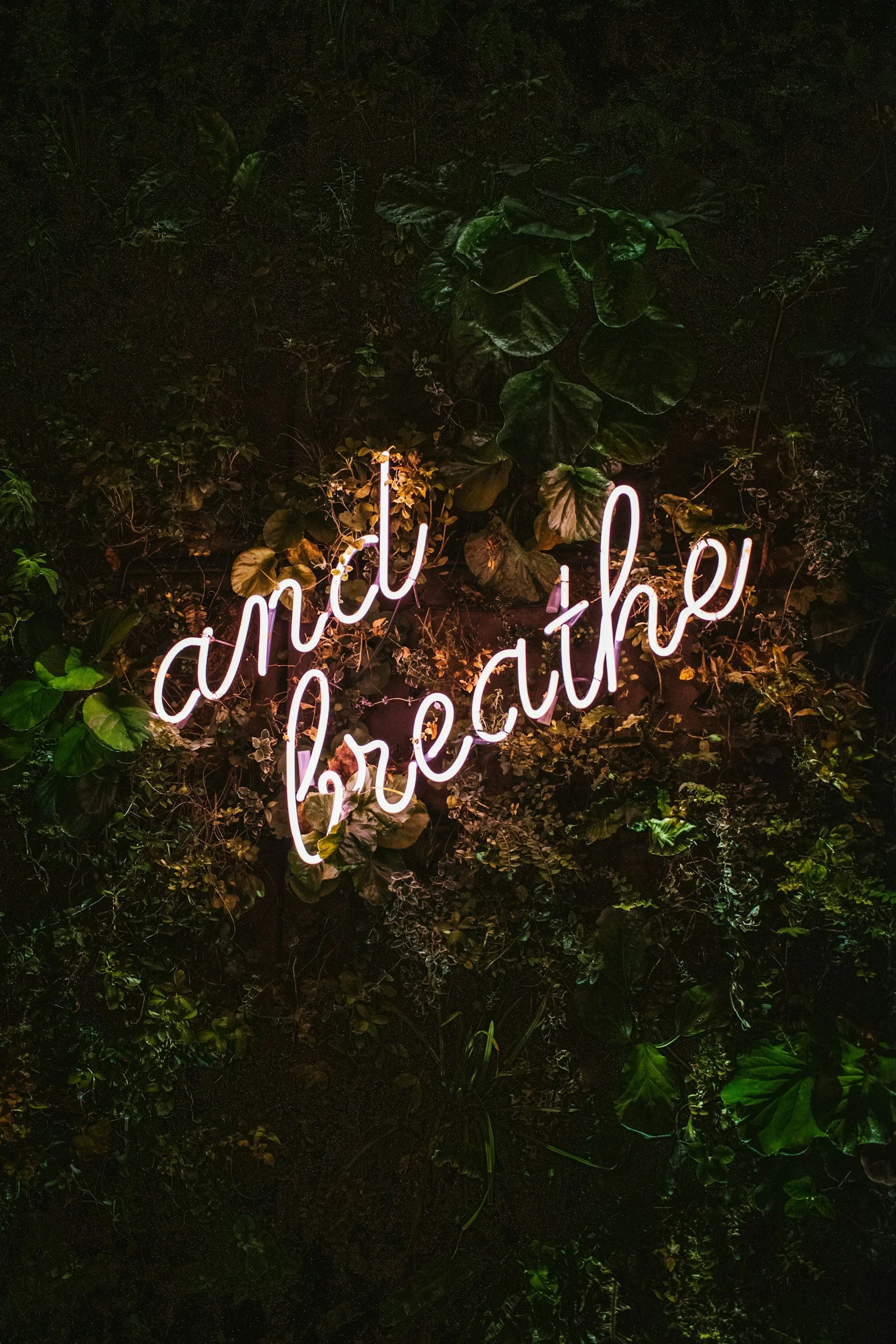 A neon sign with the words "and I live for the" hangs over a lush green wall of plants and leaves.