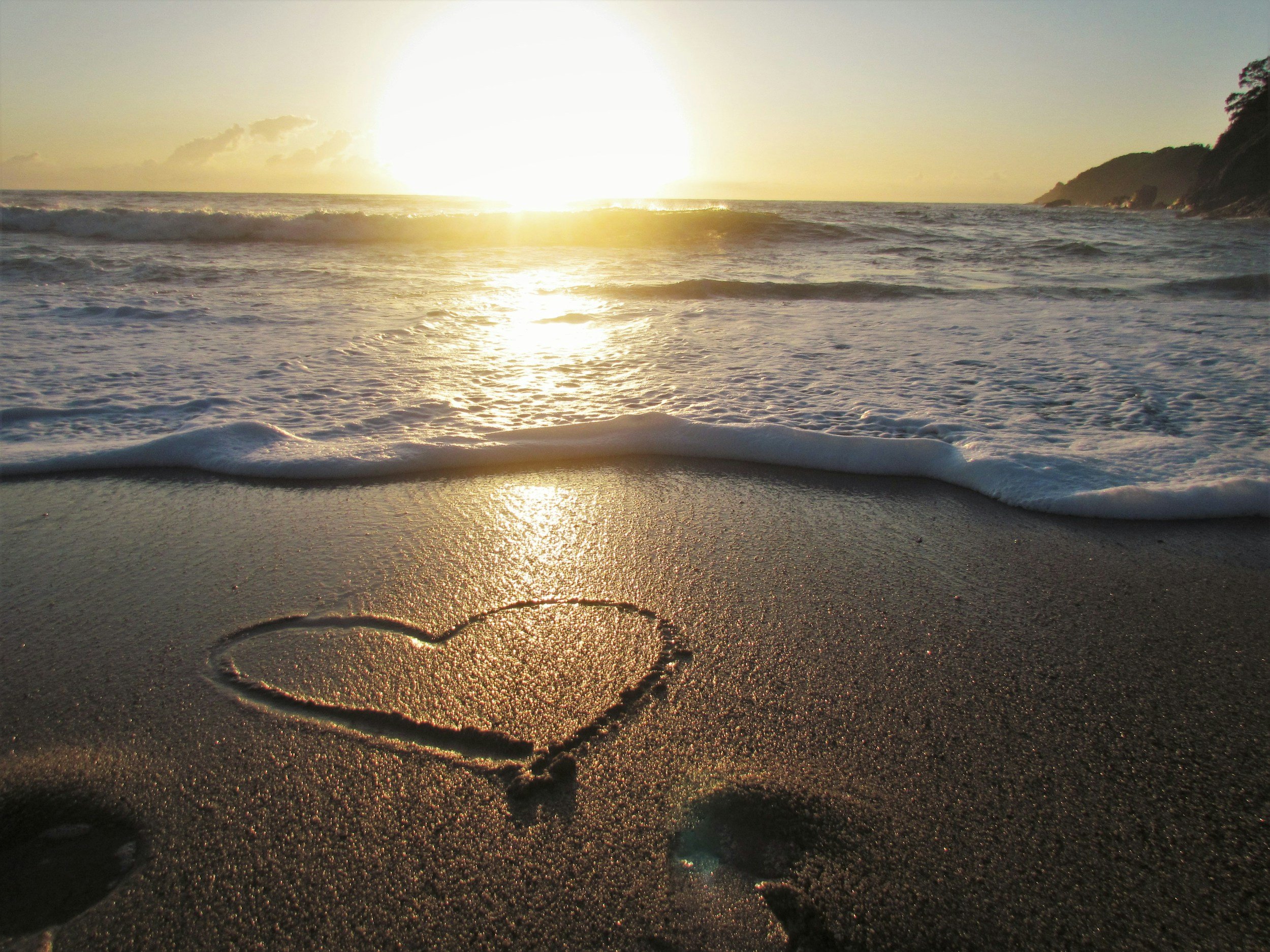 Beach scene with the sun setting over the ocean, waves crashing onto the sandy shore, and a heart shape drawn in the sand.