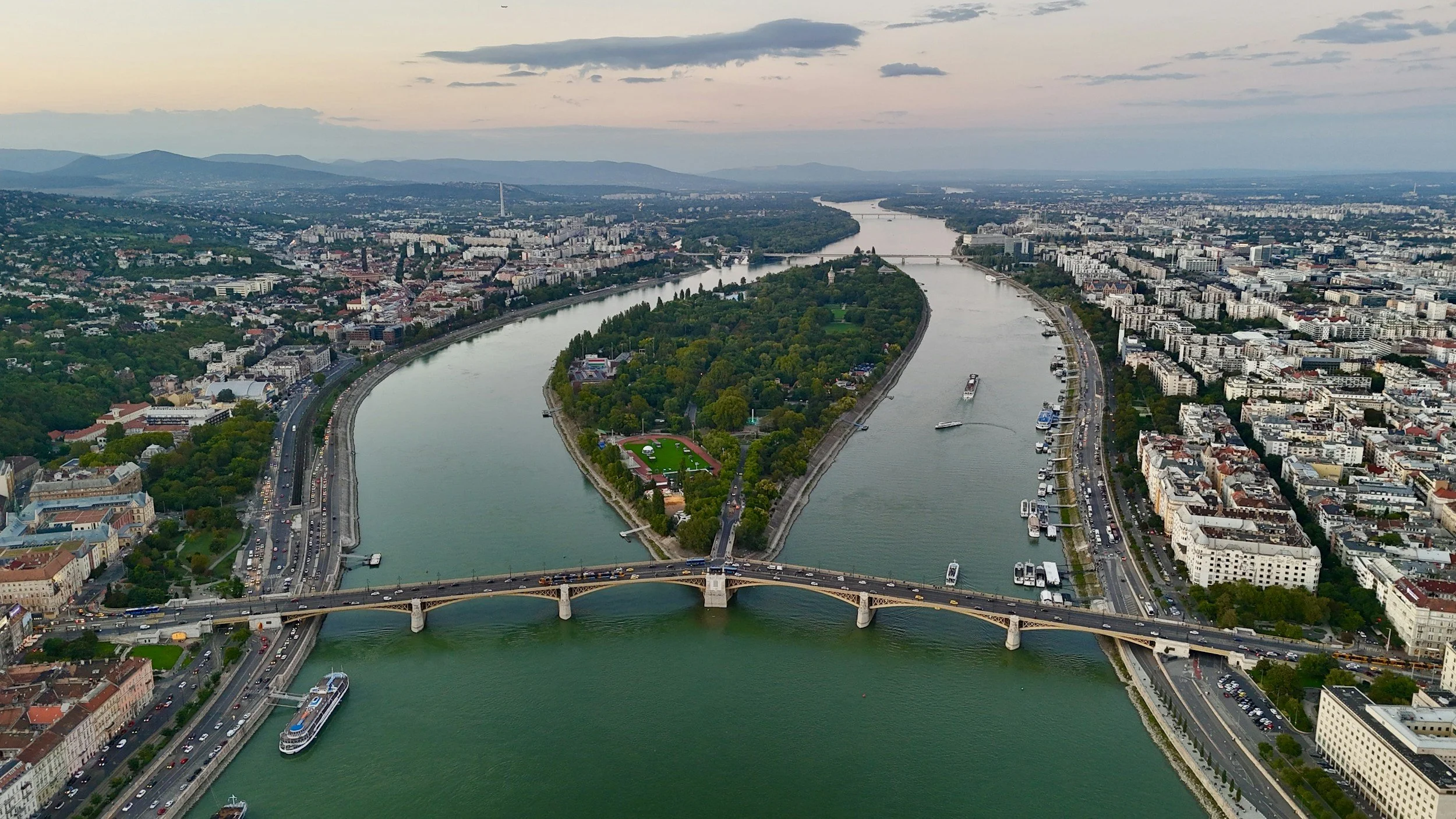 Aerial view of a city with a river running through the center, multiple bridges crossing the river, and a mix of urban and green areas during sunset.