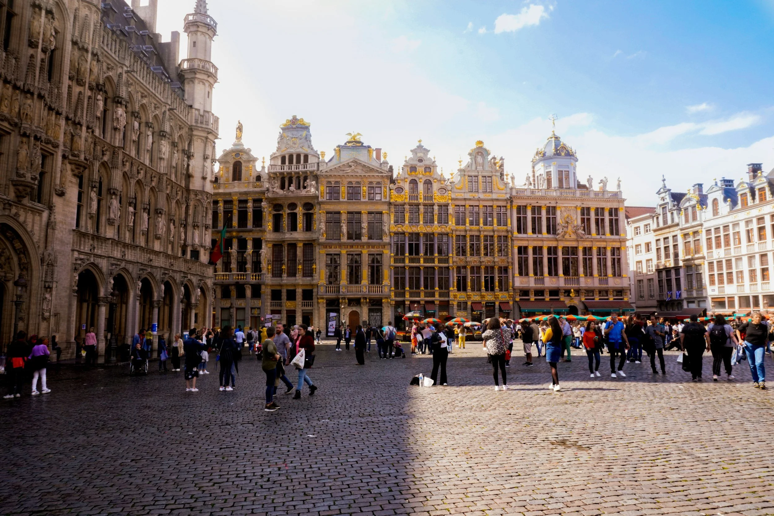 People walking and gathering in a historic European city square with ornate buildings and cobblestone pavement.