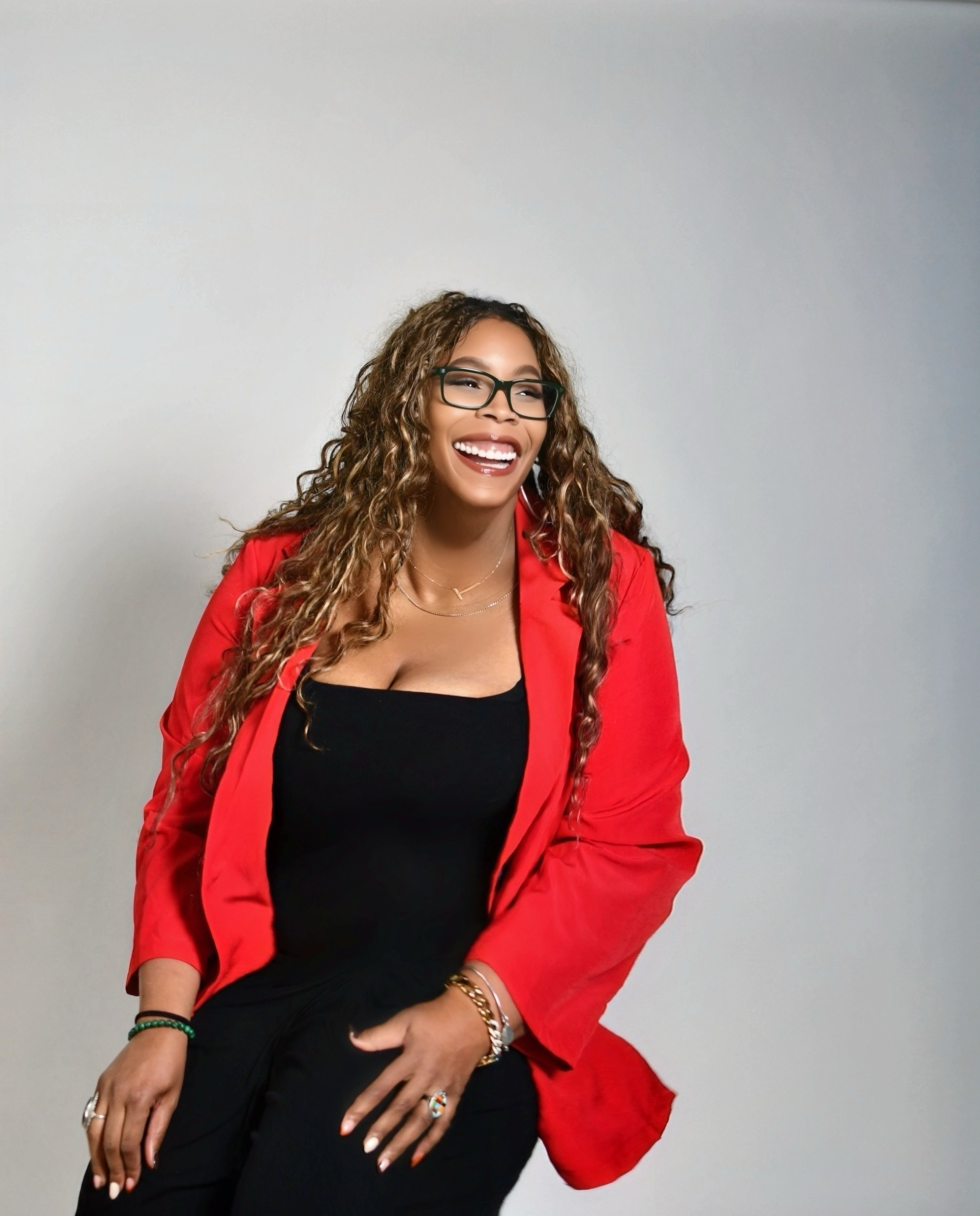 A woman with long, curly hair wearing a red blazer and black pants, sitting on a mustard-colored sofa in a room with wooden wall paneling, looking to the side.