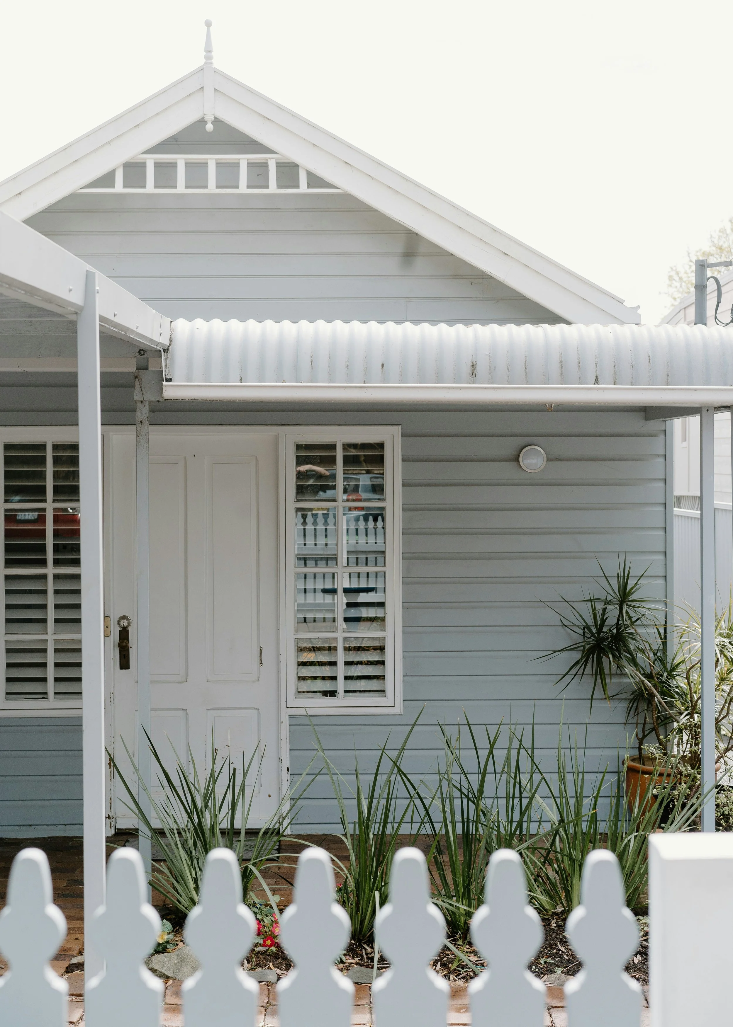 Front view of a white house with gray siding, a white door, a window with white shutters, potted plants, a small garden, and a white picket fence.