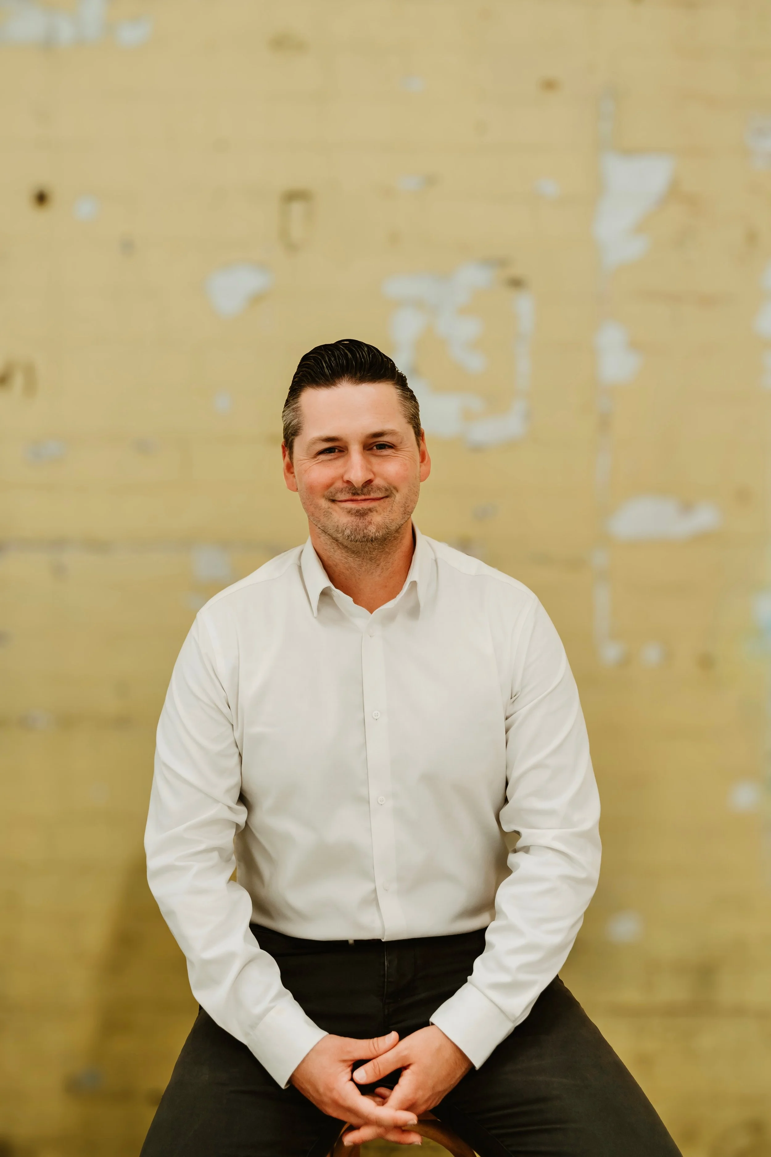 A man with dark, slicked-back hair, wearing a white button-down shirt and black pants, sitting on a stool against a yellowish, textured wall with peeling paint.