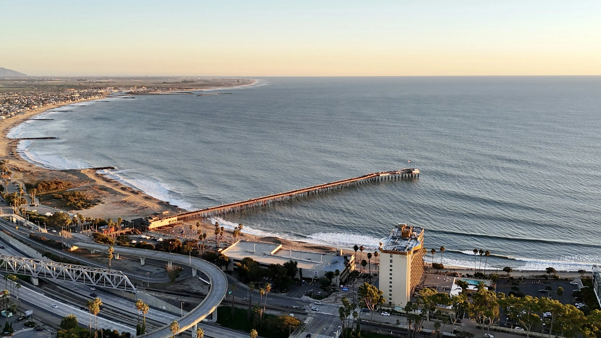 Aerial view of a beach with a pier extending into the ocean, surrounded by a coastal city with buildings, roads, and palm trees.