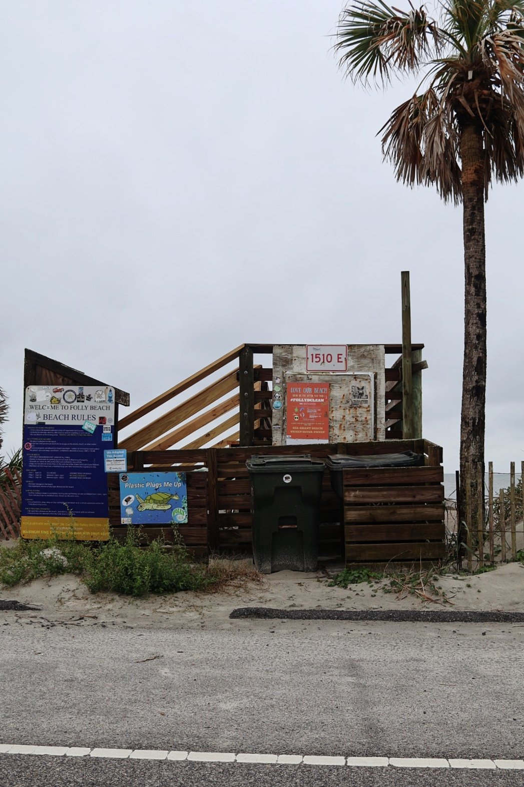 Beach access sign with warning and rules, trash bins, some greenery, palm tree, and cloudy sky at Folly Beach.