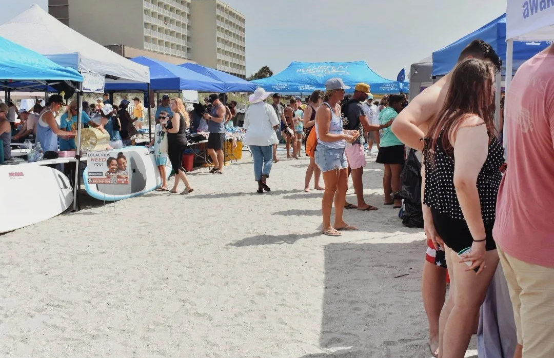 People standing in line at outdoor market stalls with blue and white canopies on the beach, some wearing hats and casual summer clothing.