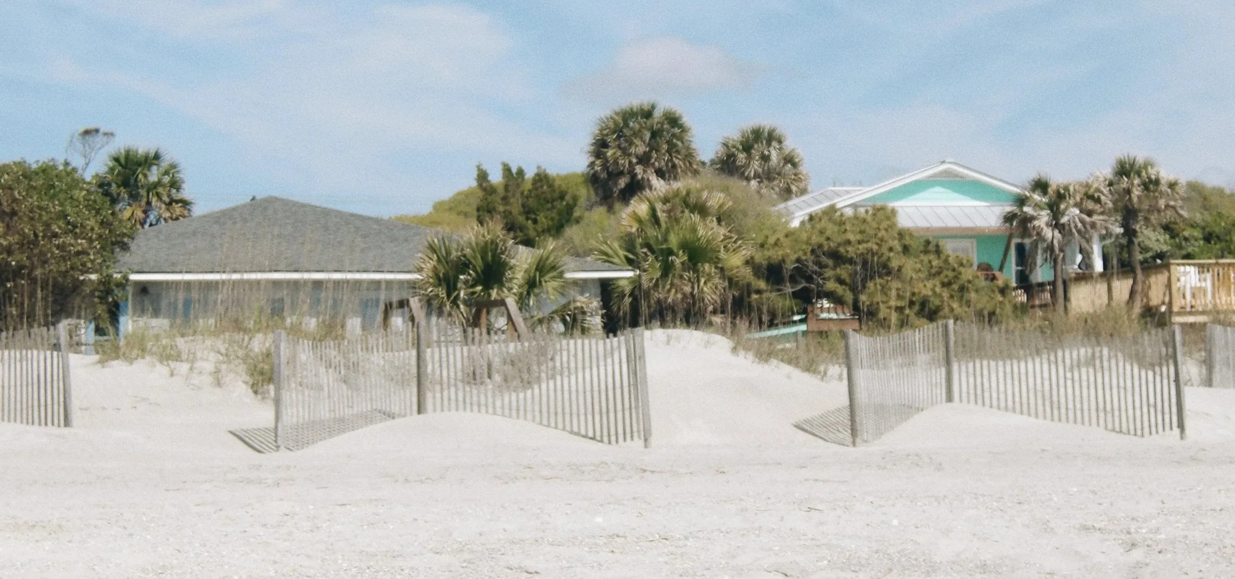 A sandy beach with a wooden fence, palm trees, and colorful houses in the background under a partly cloudy sky.