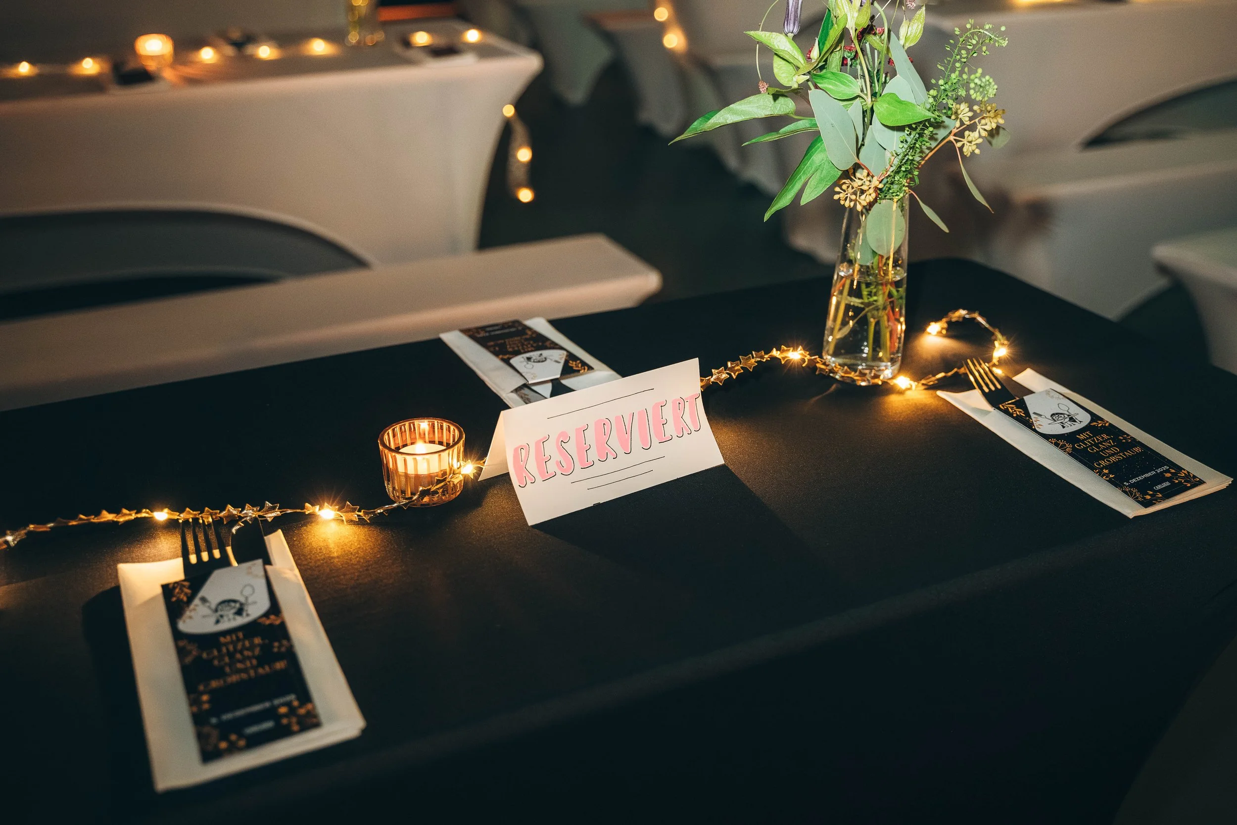 Ein festlich gedeckter Tisch mit einem Blumenstrauß, einer Kerze, einer reserviert-Karte, Besteck und Papptellern in einer eleganten Umgebung, wahrscheinlich für eine Veranstaltung oder Feier.