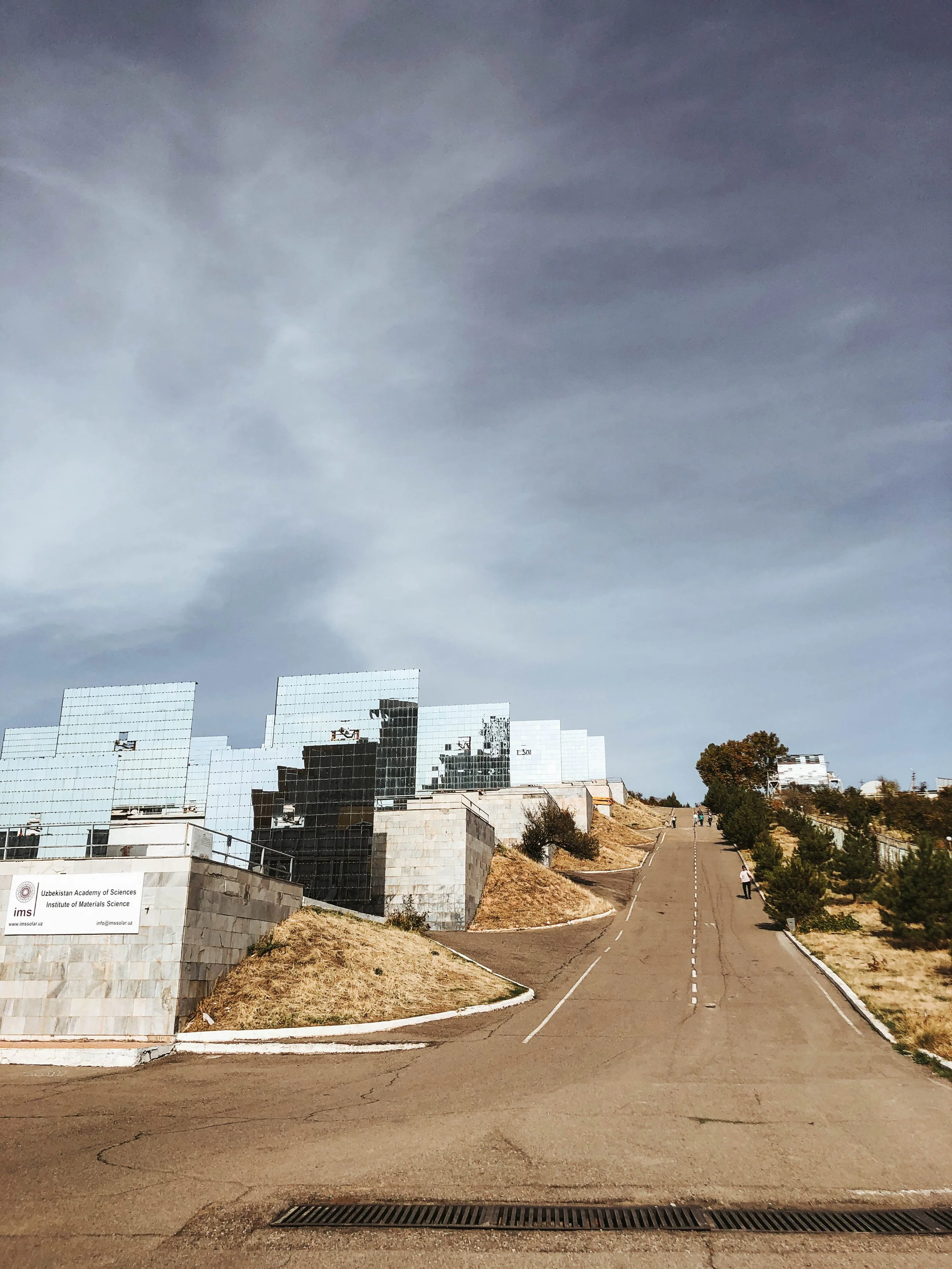 Eine steile, asphaltierte Straße führt bergauf an der Seite eines Hügels mit trockener Vegetation, links befindet sich das Gebäude der Usbekischen Akademie der Wissenschaften mit Glasfassade, im Hintergrund sind ein paar Menschen zu sehen