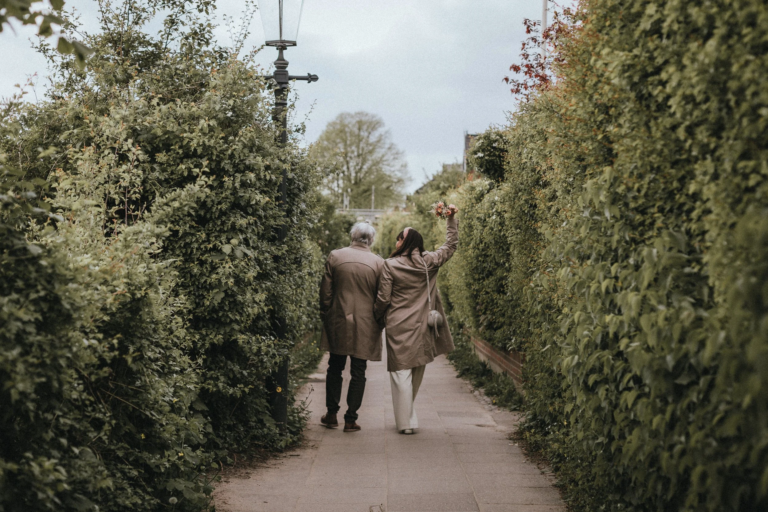 Ein Paar geht Hand in Hand durch einen grünen Gehweg mit hohen Hecken auf beiden Seiten, eine Frau hält einen Blumenstrauß in der Hand.