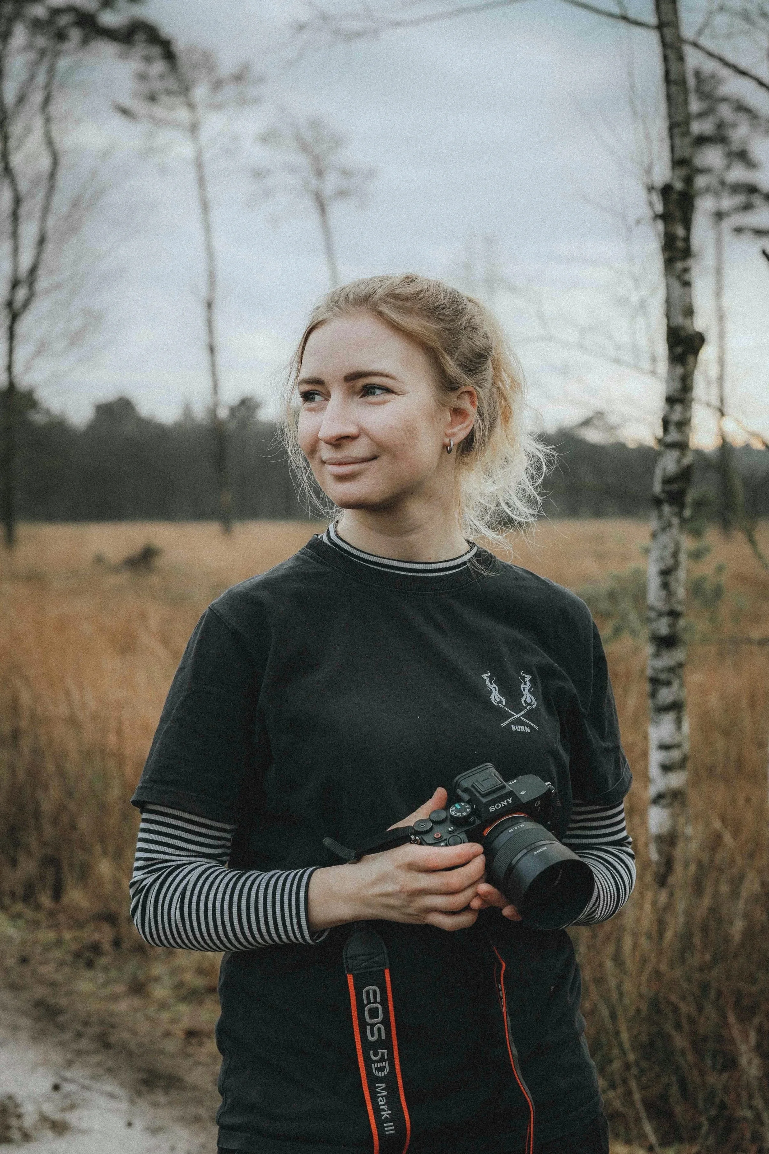 Eine junge Frau mit blondem Haar, die draußen in einem Feld steht und eine Kamera hält.