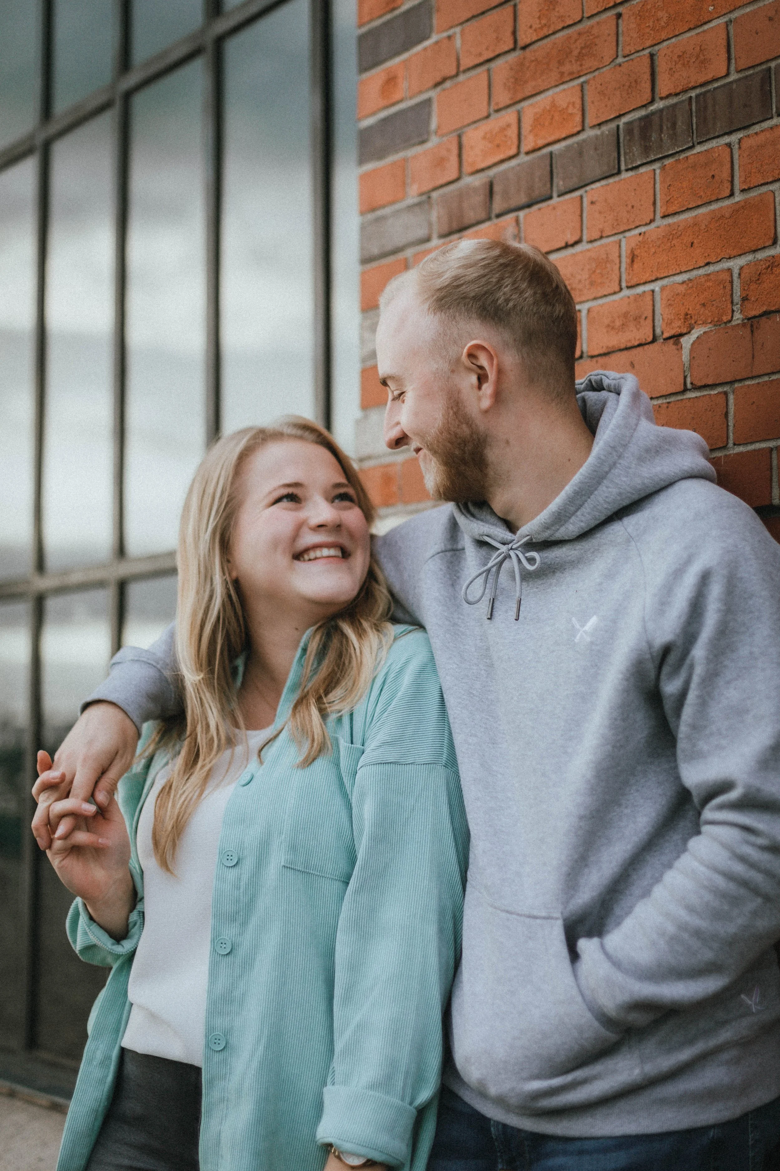 Ein lächelndes junges Paar steht nebeneinander vor einem roten Backsteinmauer und einer großen Fensterwand, umarmt sich und schaut sich liebevoll an.