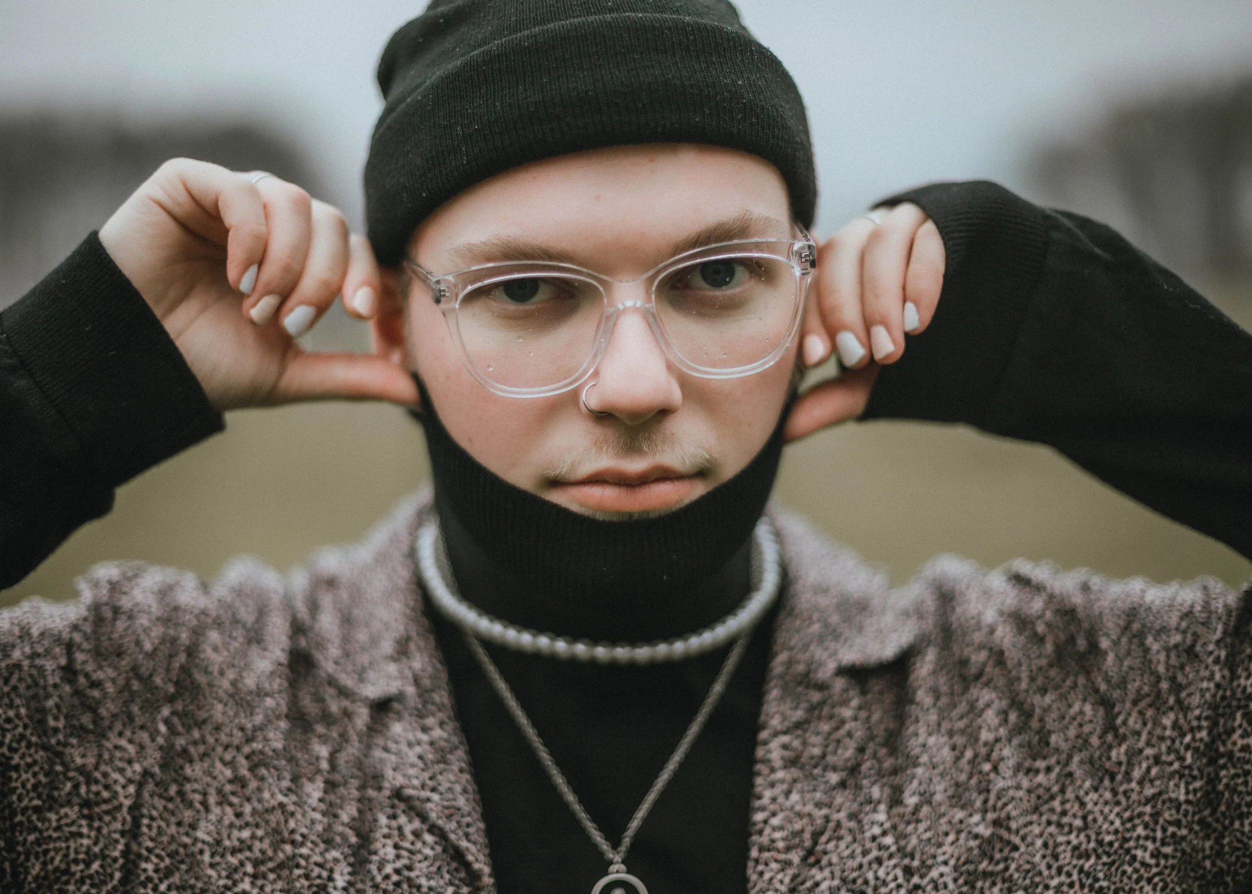 Nahaufnahme eines jungen Erwachsenen mit Brille, schwarzem Tuch um den Hals, Mütze, und mehreren Halsketten. Der Hintergrund ist unscharf, vermutlich ein freier, bewölkter Himmel.