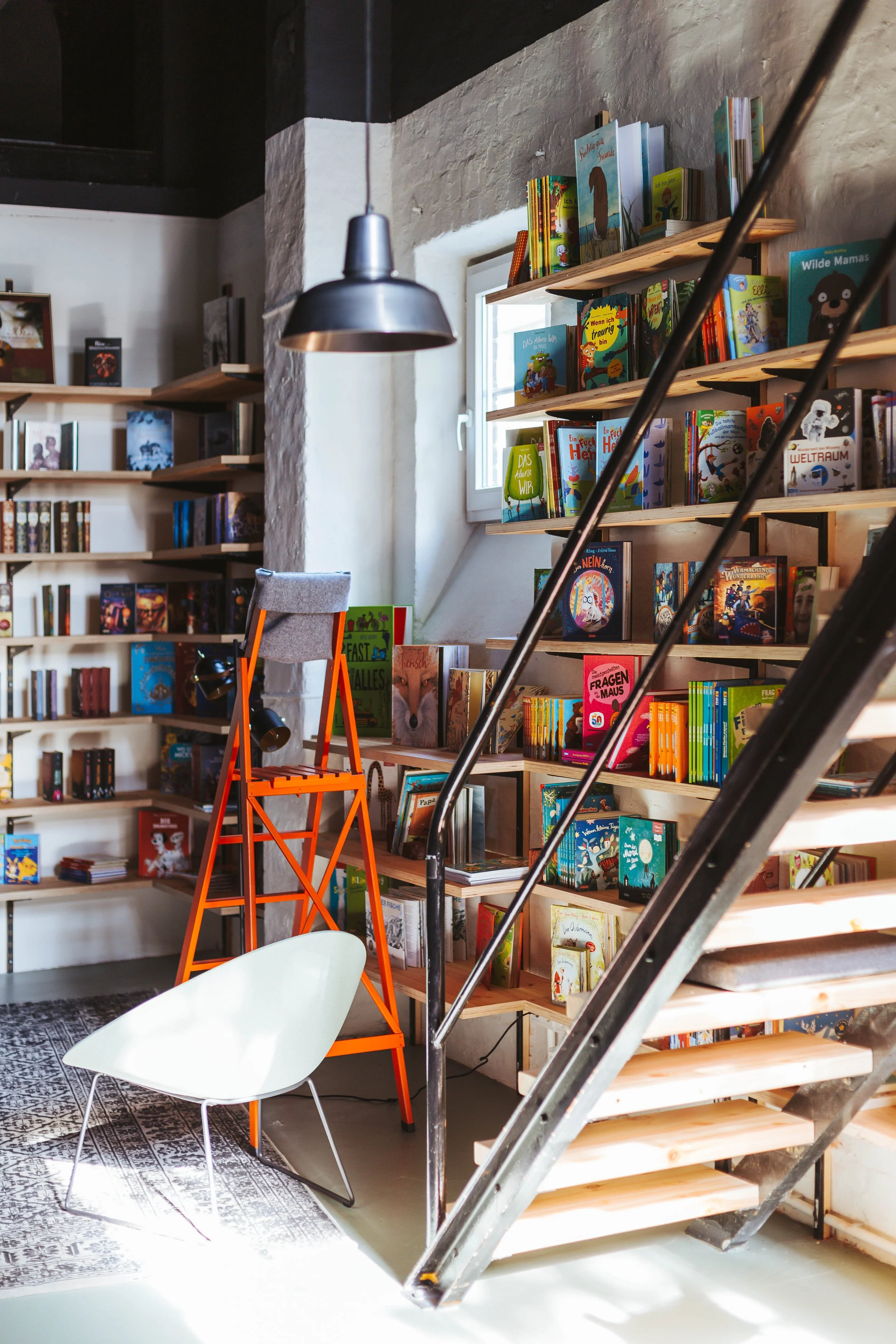 Buchhandlung mit Holzregalen voller bunter Kinderbücher, eine orange Leiter, ein weißer Stuhl, ein Fenster und eine helle Beleuchtung.