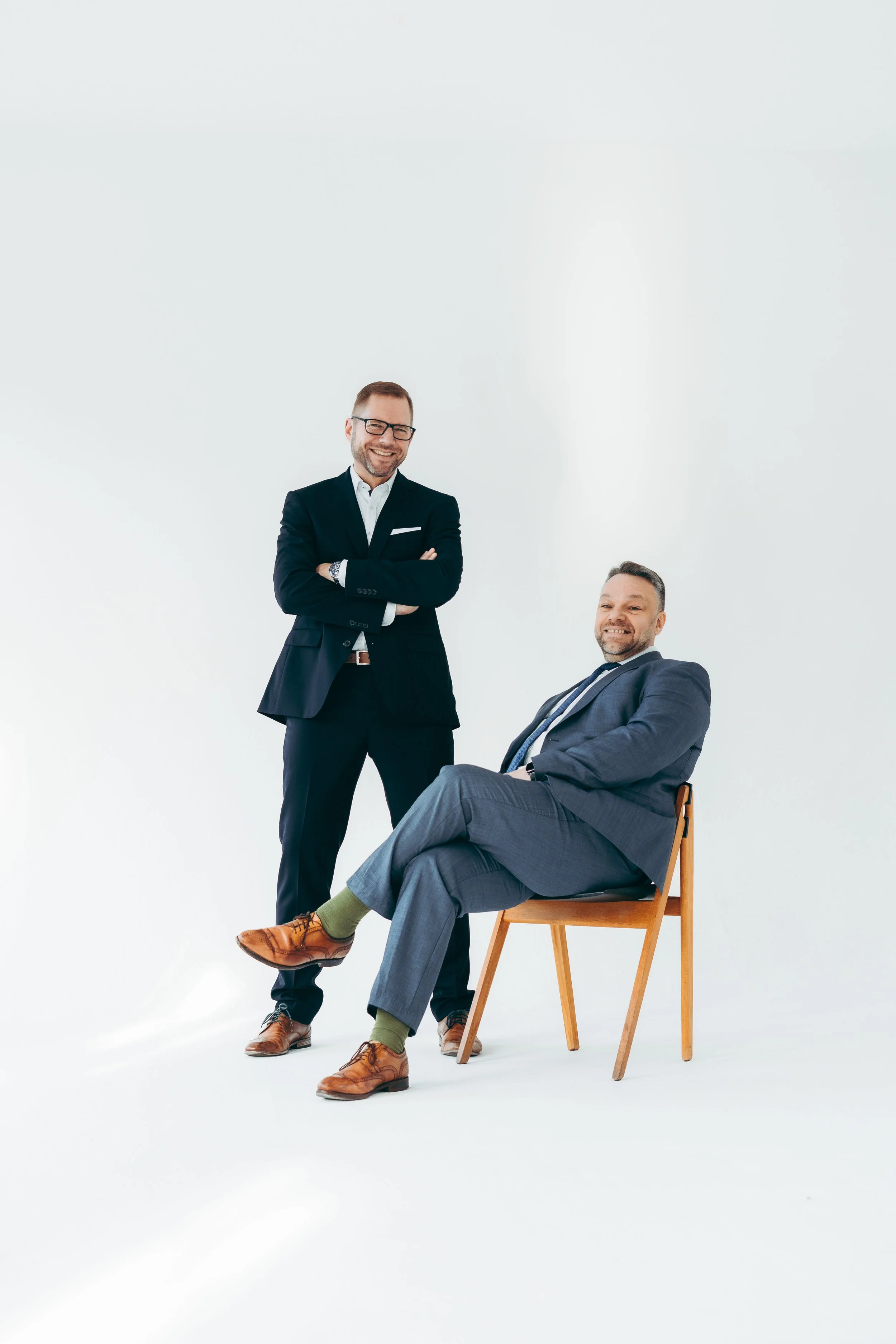 Zwei Männer in Anzügen posieren vor weißem Hintergrund, einer sitzt auf einem Stuhl und der andere steht daneben.