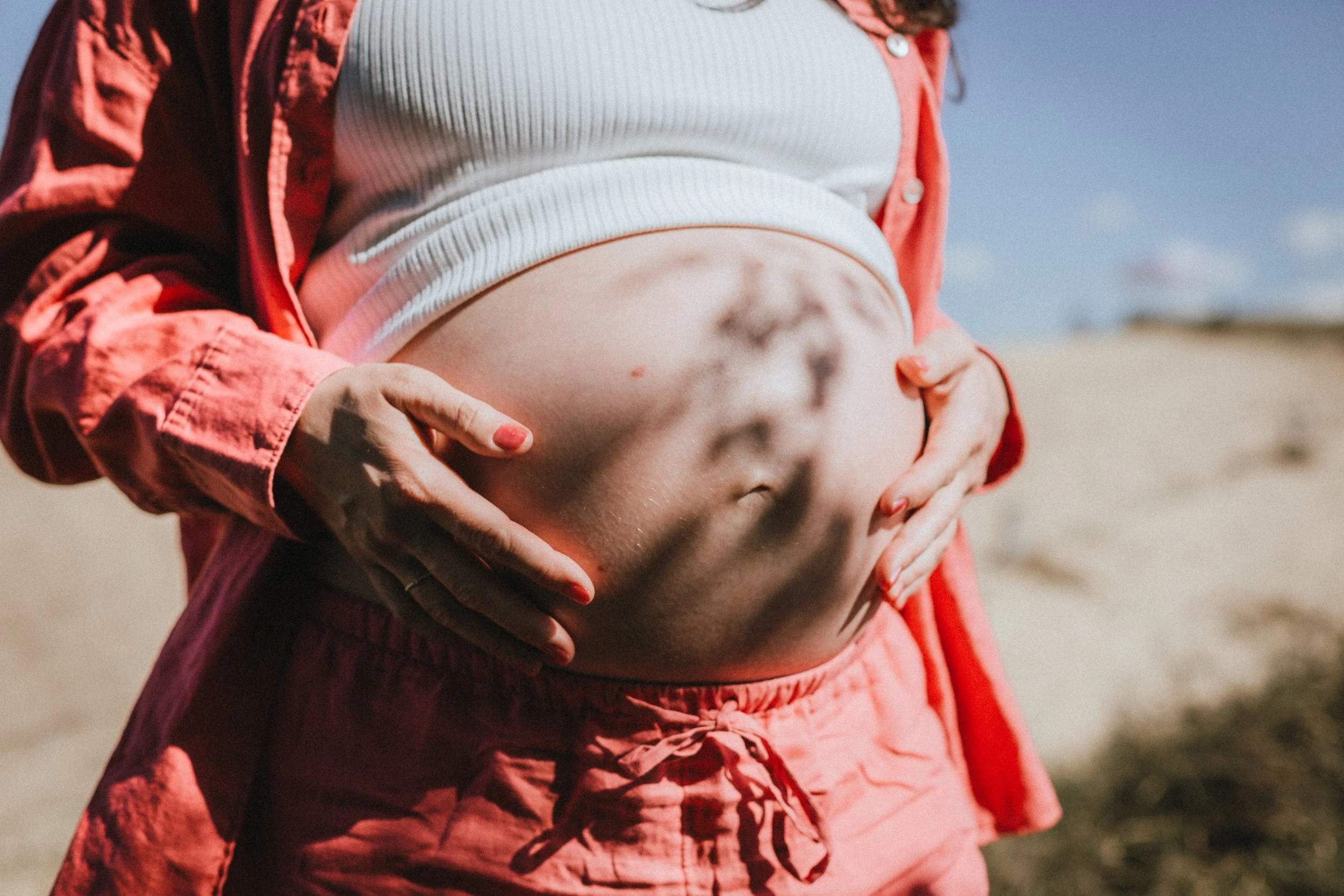 Nahaufnahme eines schwangeren Bauchs, von einer Person mit roten Shorts und einer weißen, gestreiften Oberseite, die sich am Bauch berührt, im Freien bei sonnigem Wetter.