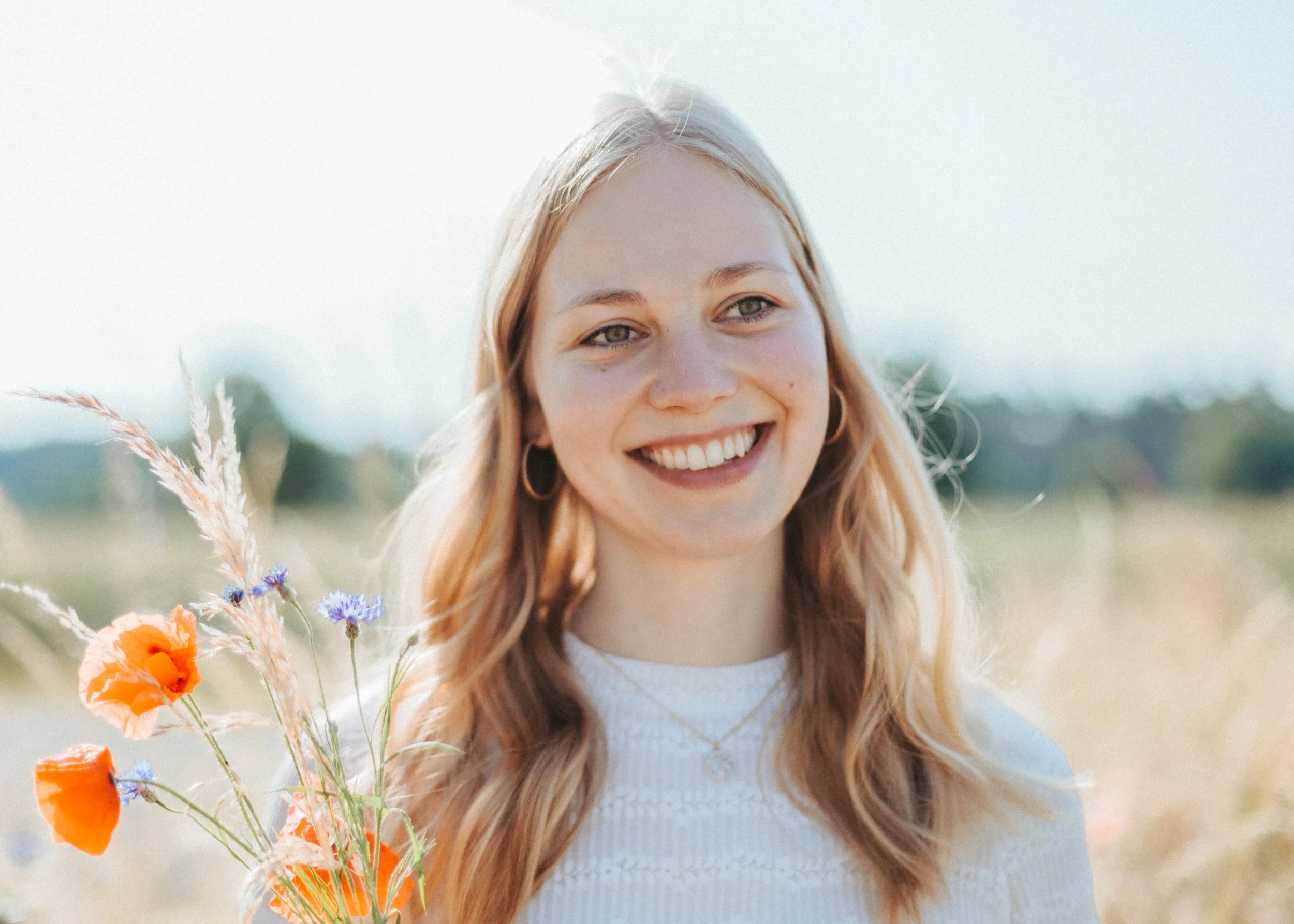 Lächelnde junge Frau hält einen Blumenstrauß aus Mohn und Kornblumen in einem sonnigen Feld.