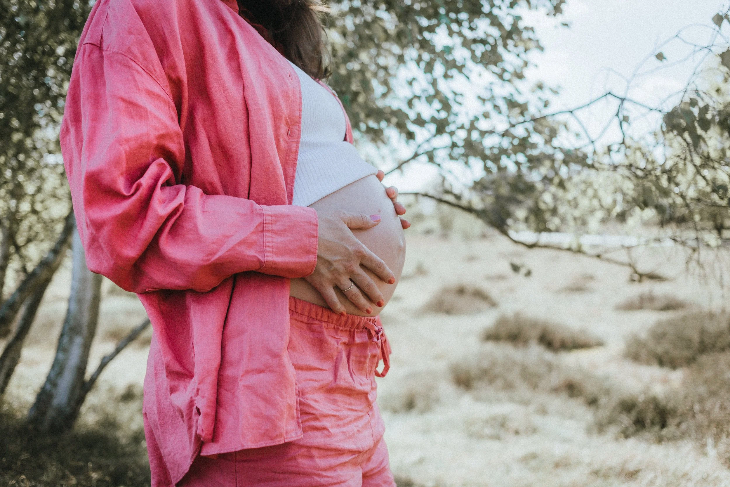 Eine schwangere Frau, die ihren Bauch hält, steht im Freien bei Bäumen und Sonnenlicht. Sie trägt eine weiße Top und eine pinkfarbene Jacke und Hosen.
