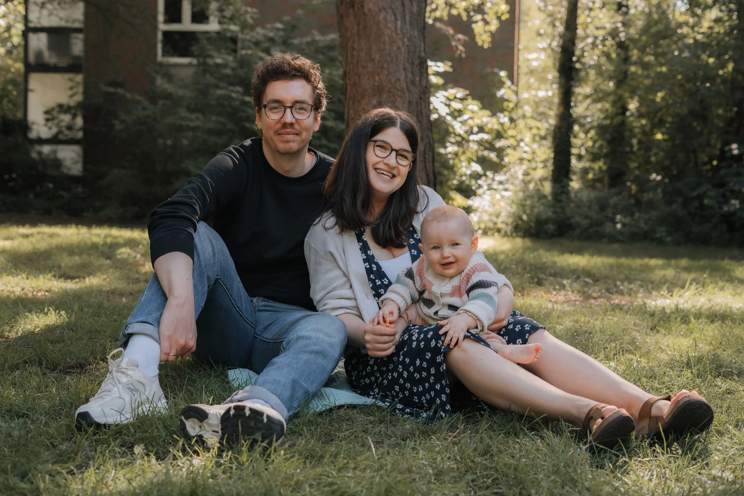Familie mit einem Vater, einer Mutter und einem Baby sitzt auf dem Gras in einem Park mit Bäumen im Hintergrund, Sonne scheint durch die Blätter.
