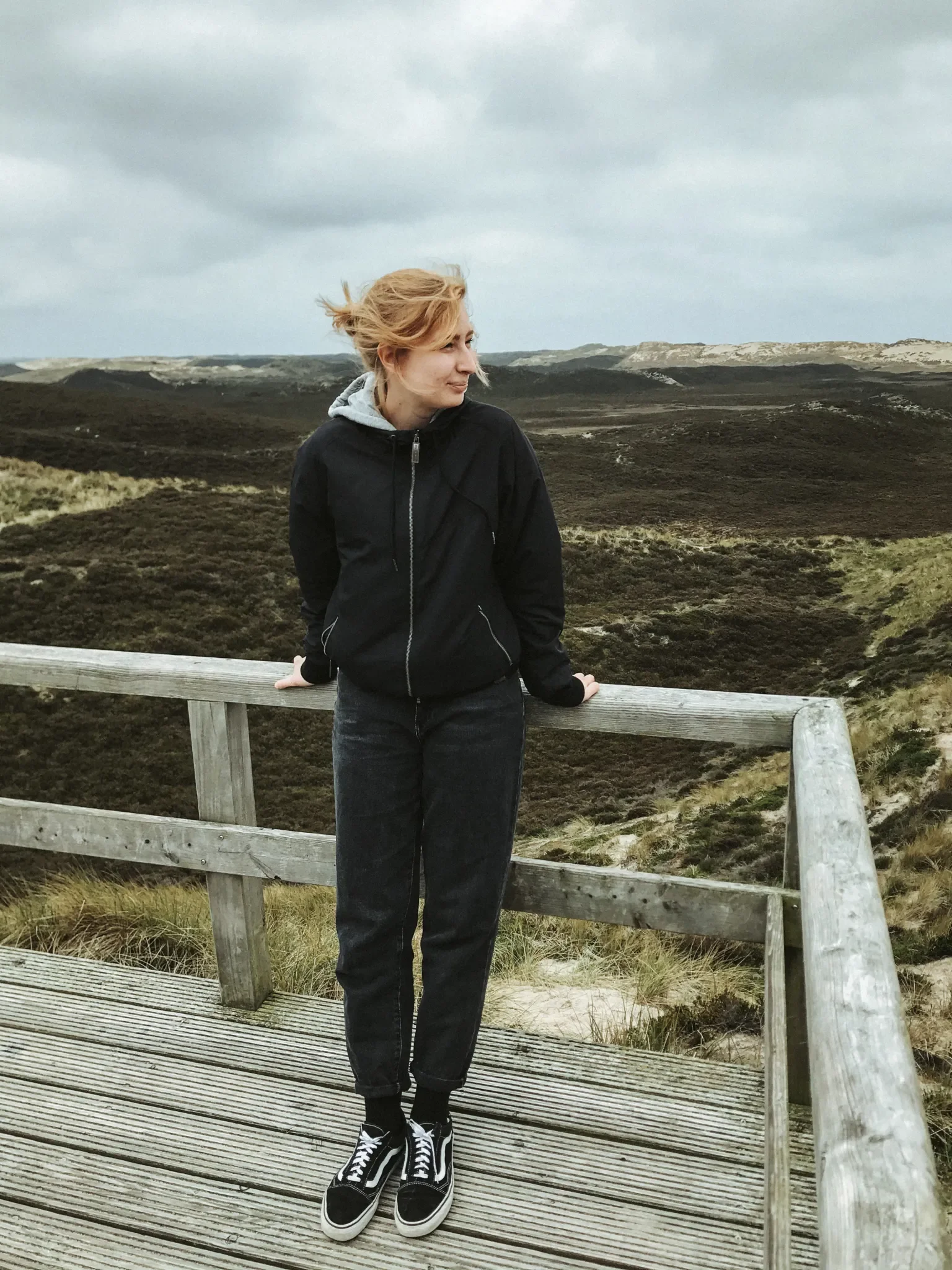 Eine junge Frau steht auf einer Holzkonstruktion inmitten einer ländlichen, bewaldeten Landschaft mit bewölktem Himmel im Hintergrund.