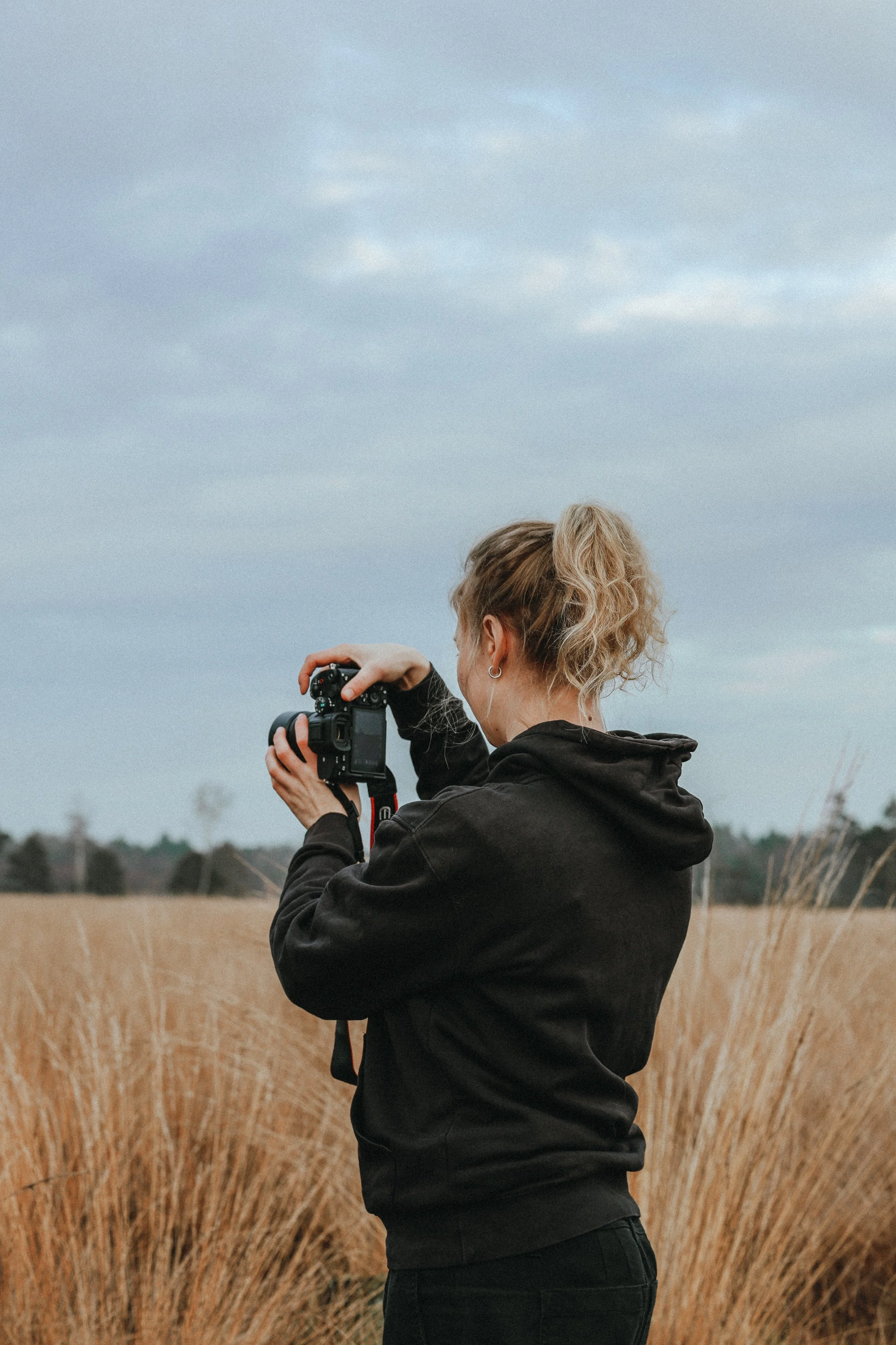 Eine junge Frau mit lockigem, blonden Haar in einem schwarzen Hoodie, die eine Kamera in einem offenen Feld fotografiert, im Hintergrund Himmel und Bäume.